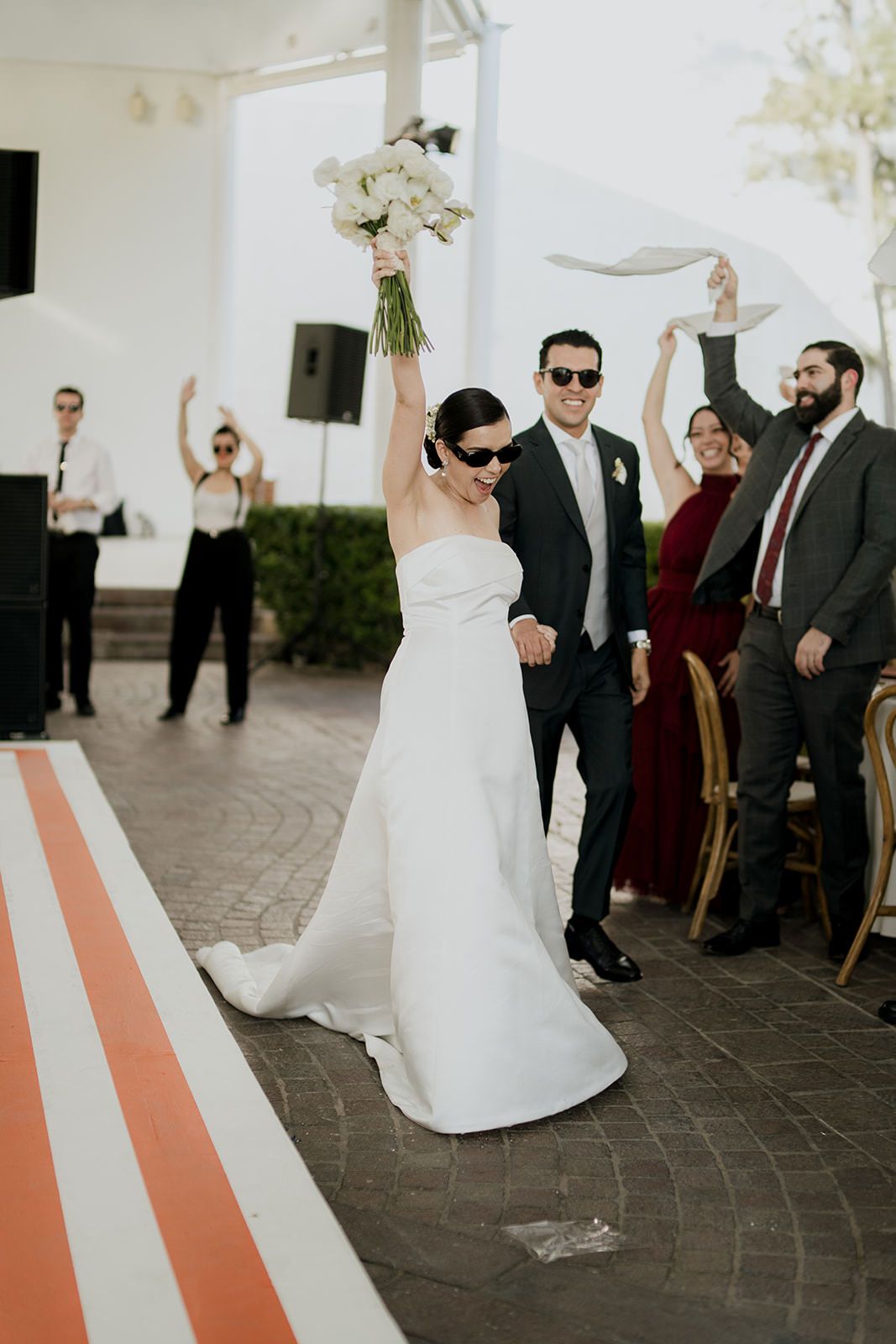 Bride raises bouquet triumphantly while groom in sunglasses walks beside her at elegant outdoor wedding reception.