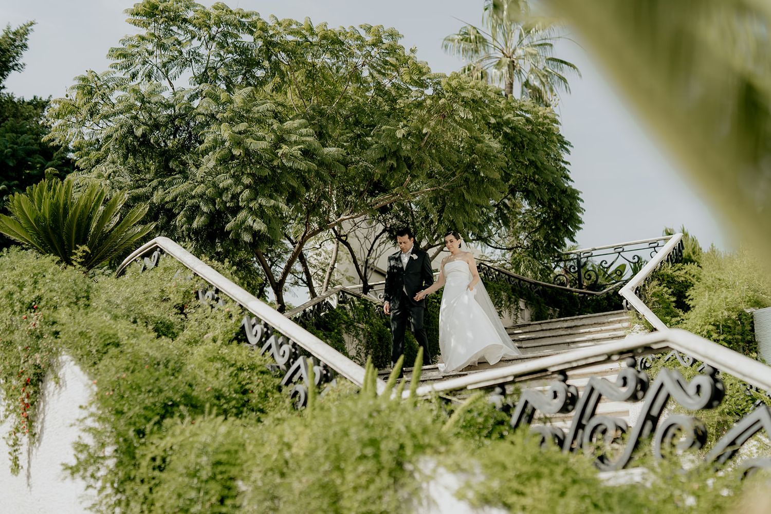 Bride and groom stand on ornate white staircase surrounded by lush tropical greenery at an outdoor wedding venue.