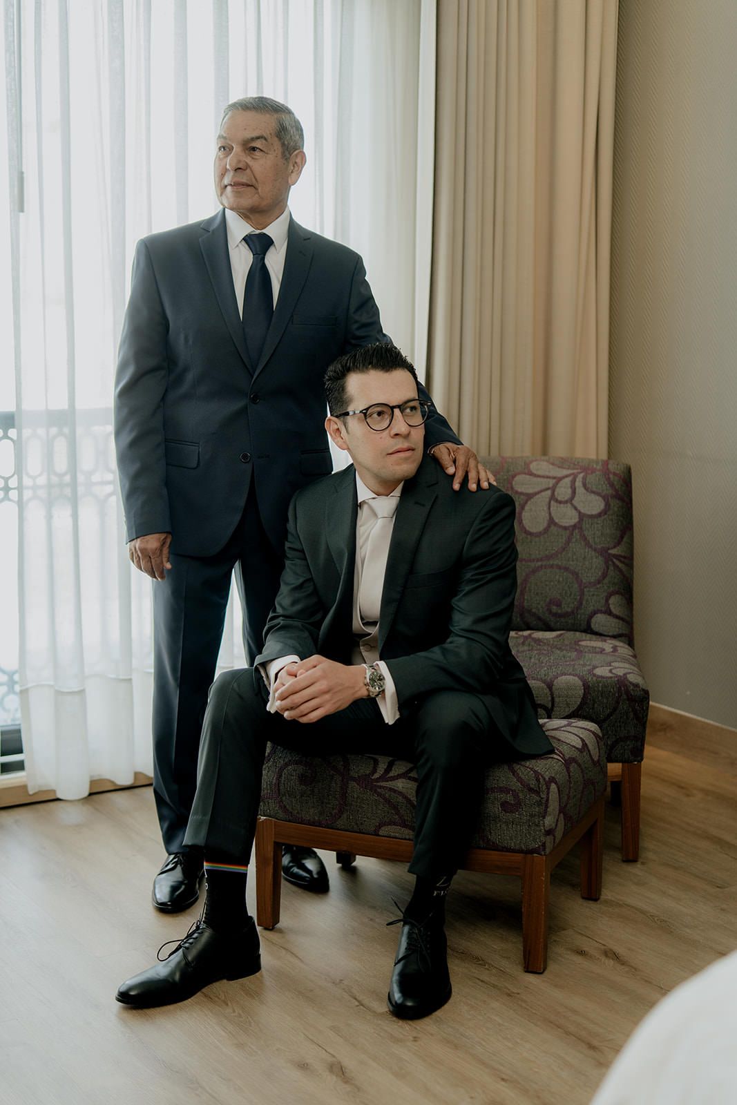 Two men in dark suits pose formally, one seated in a chair and one standing, in an elegant hotel room.