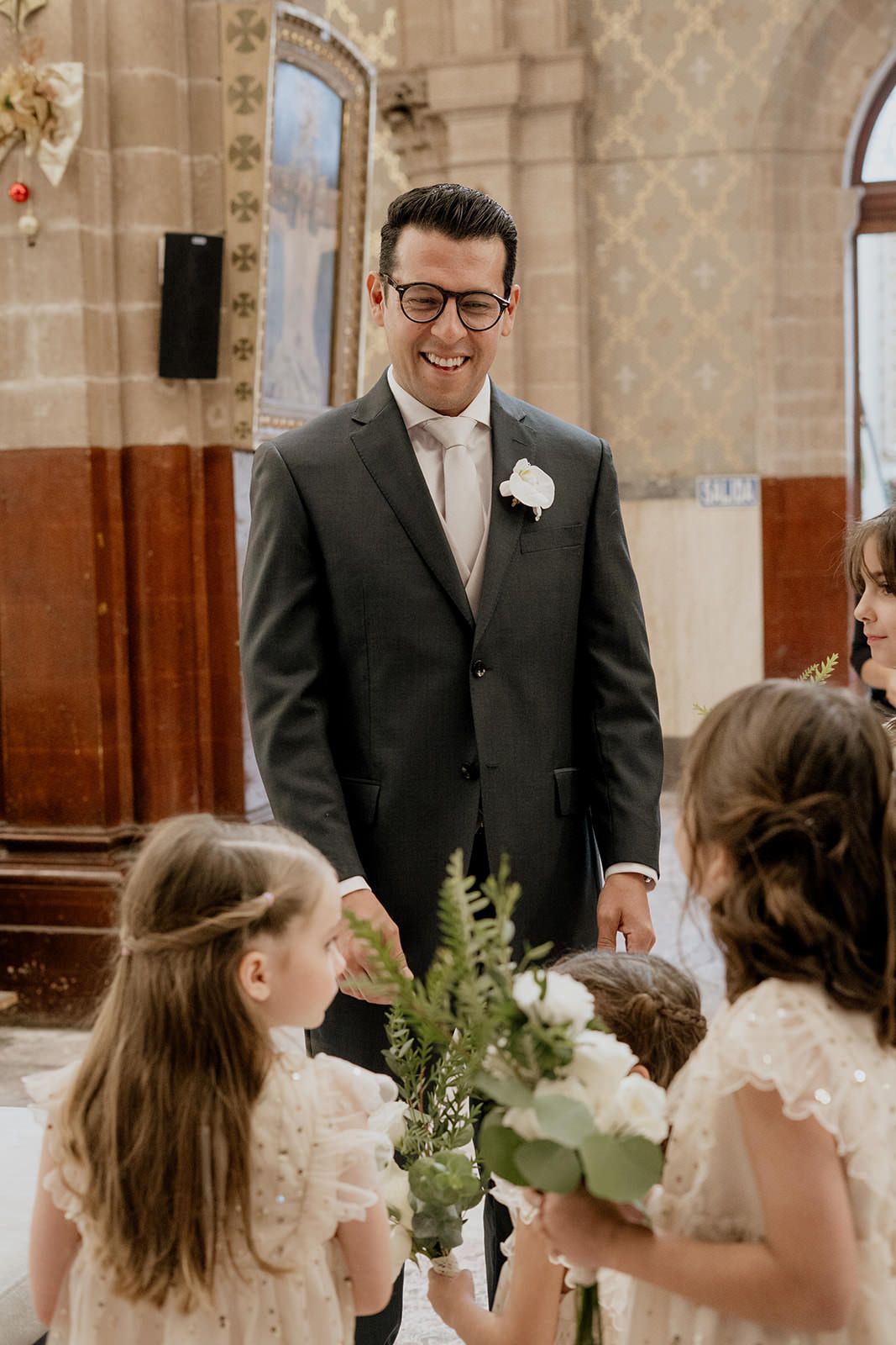 Smiling groom in dark suit with boutonniere greeting flower girls in white dresses at church wedding.