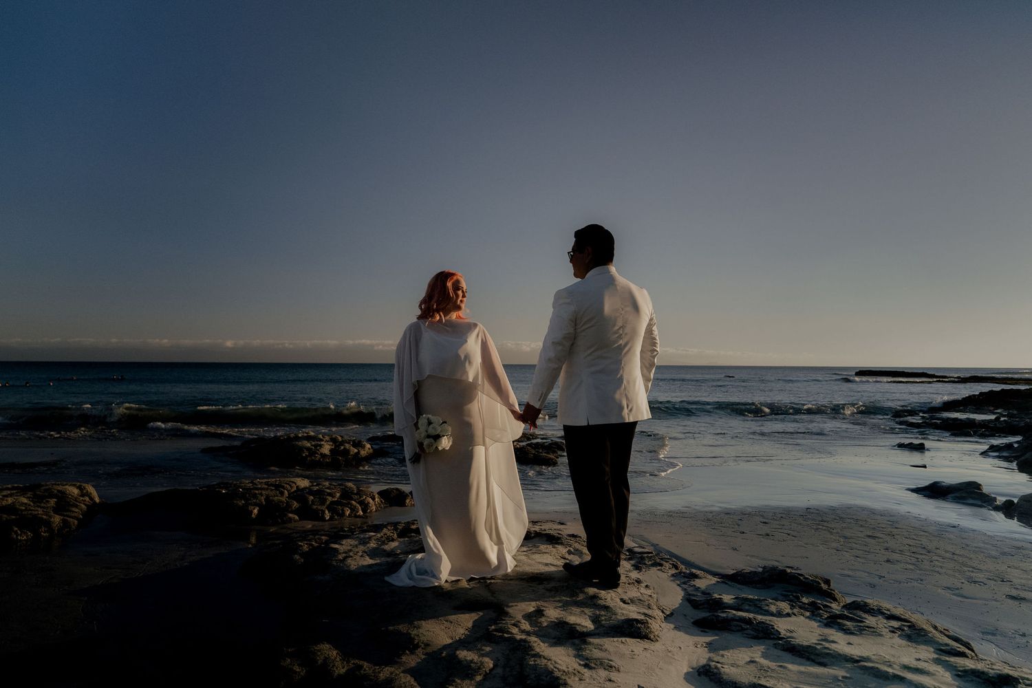 Bride and groom holding hands walking on rocky beach at sunset near ocean.