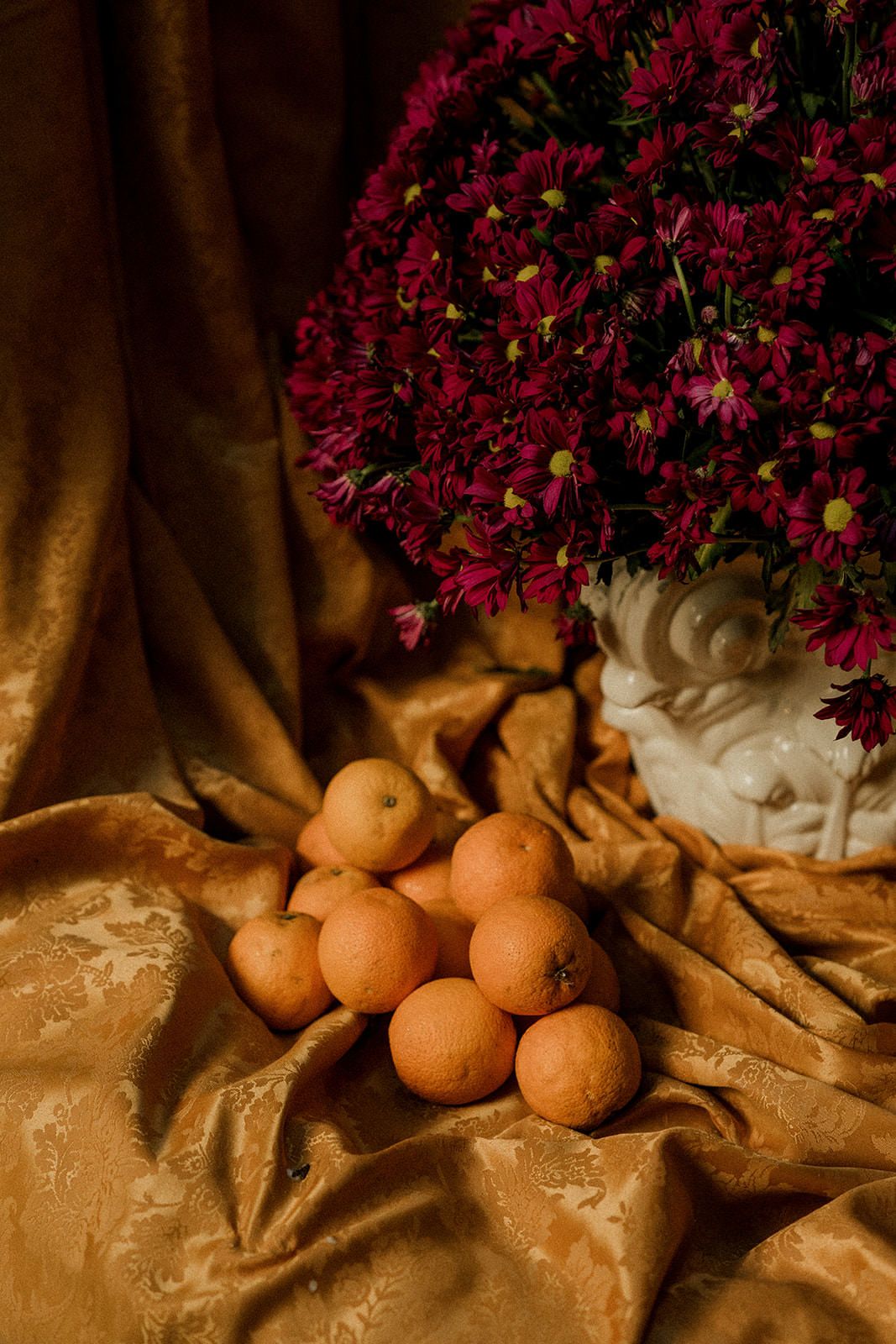 Deep burgundy chrysanthemums and fresh oranges create a dramatic still life against golden fabric in warm lighting.
