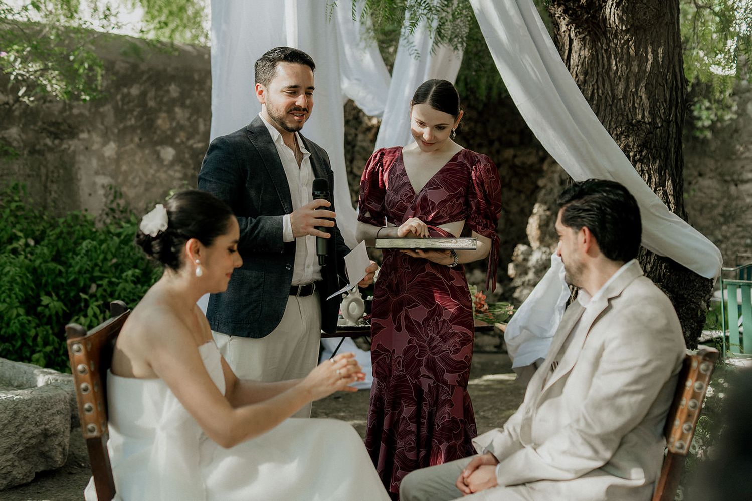 Intimate outdoor wedding ceremony under white drapes and trees with attendees gathered.