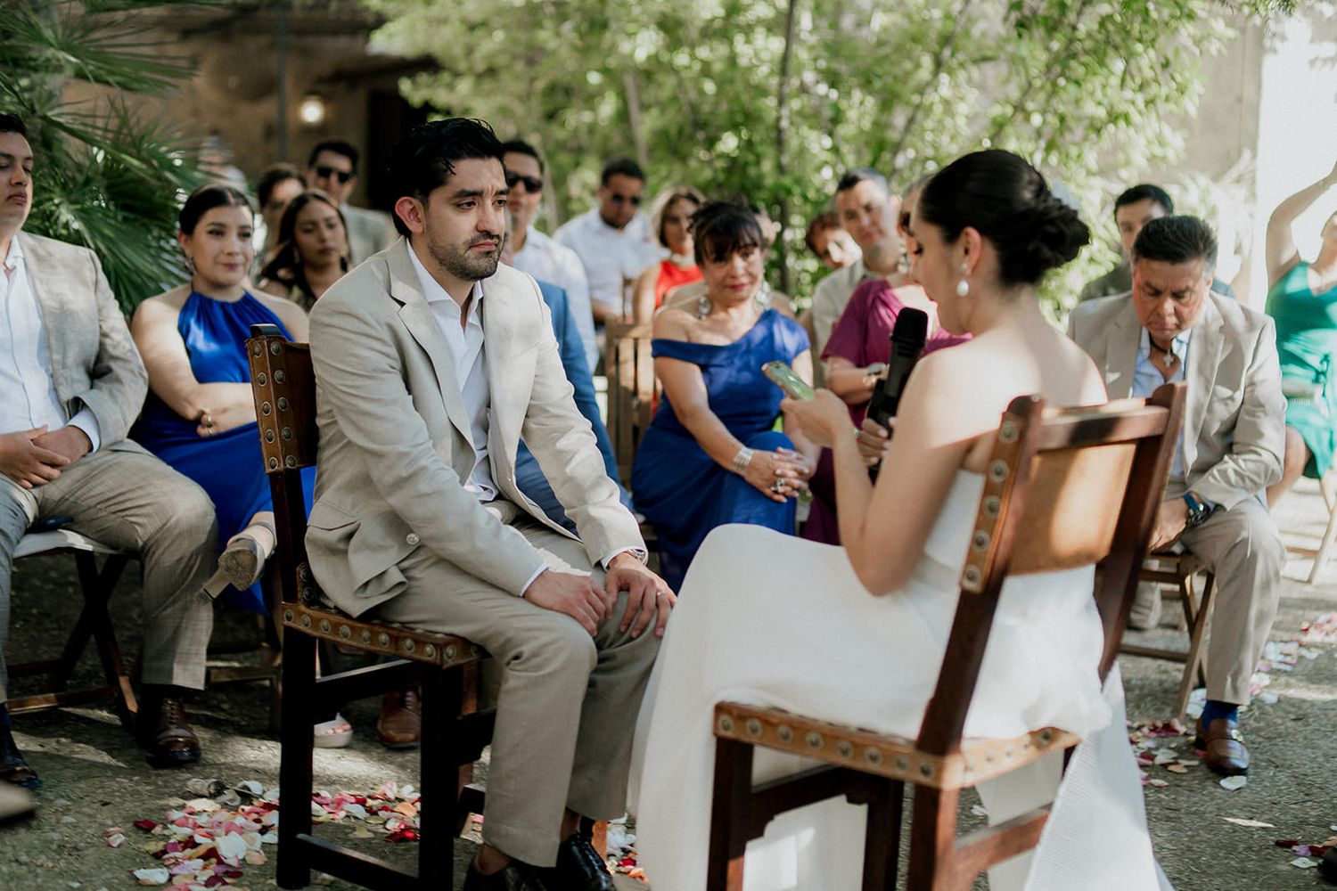 Wedding guests celebrate during an outdoor reception with white and gray attire under green trees.
