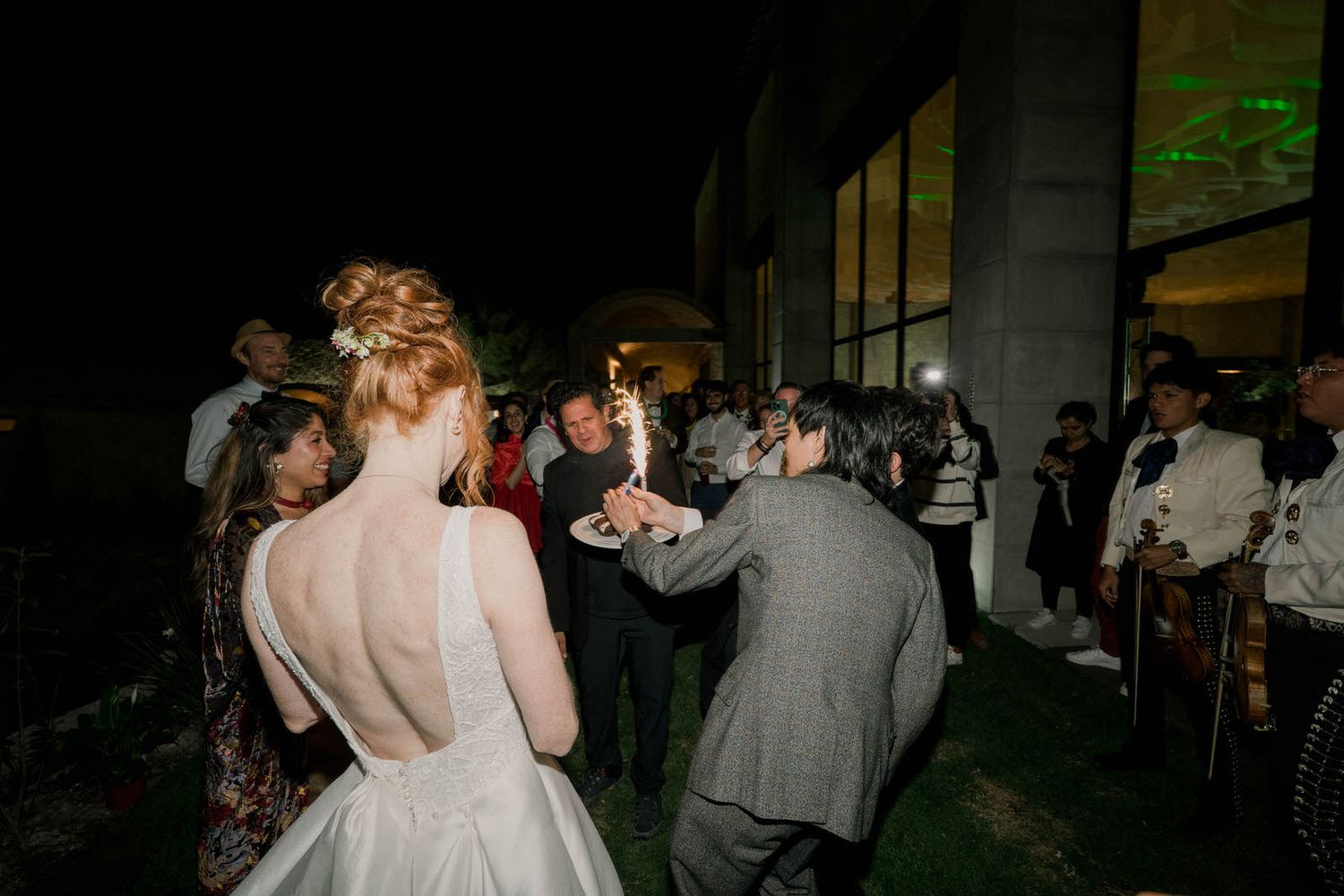 Wedding guests dance and celebrate on a dimly lit outdoor patio in the evening.