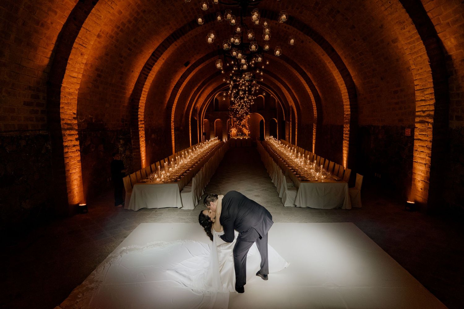 Long candlelit dining tables line a dramatic brick tunnel venue with warm ambient lighting.