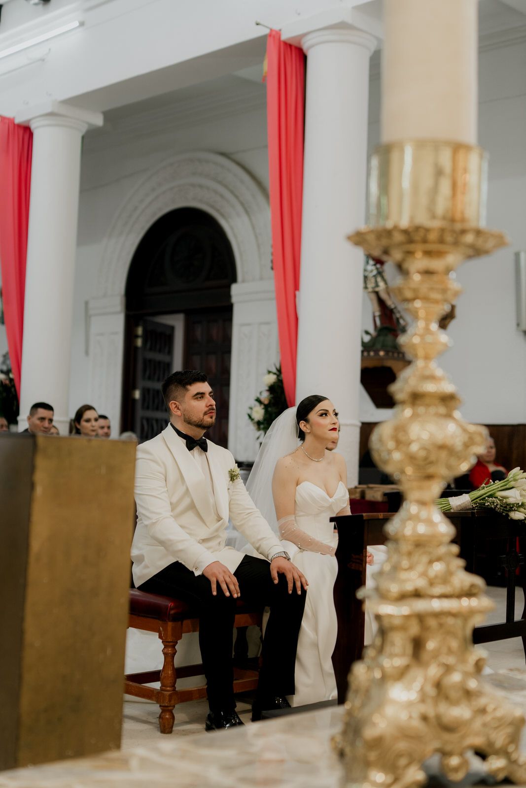 A couple sits in formal attire during their wedding ceremony with a golden candlestick in foreground.