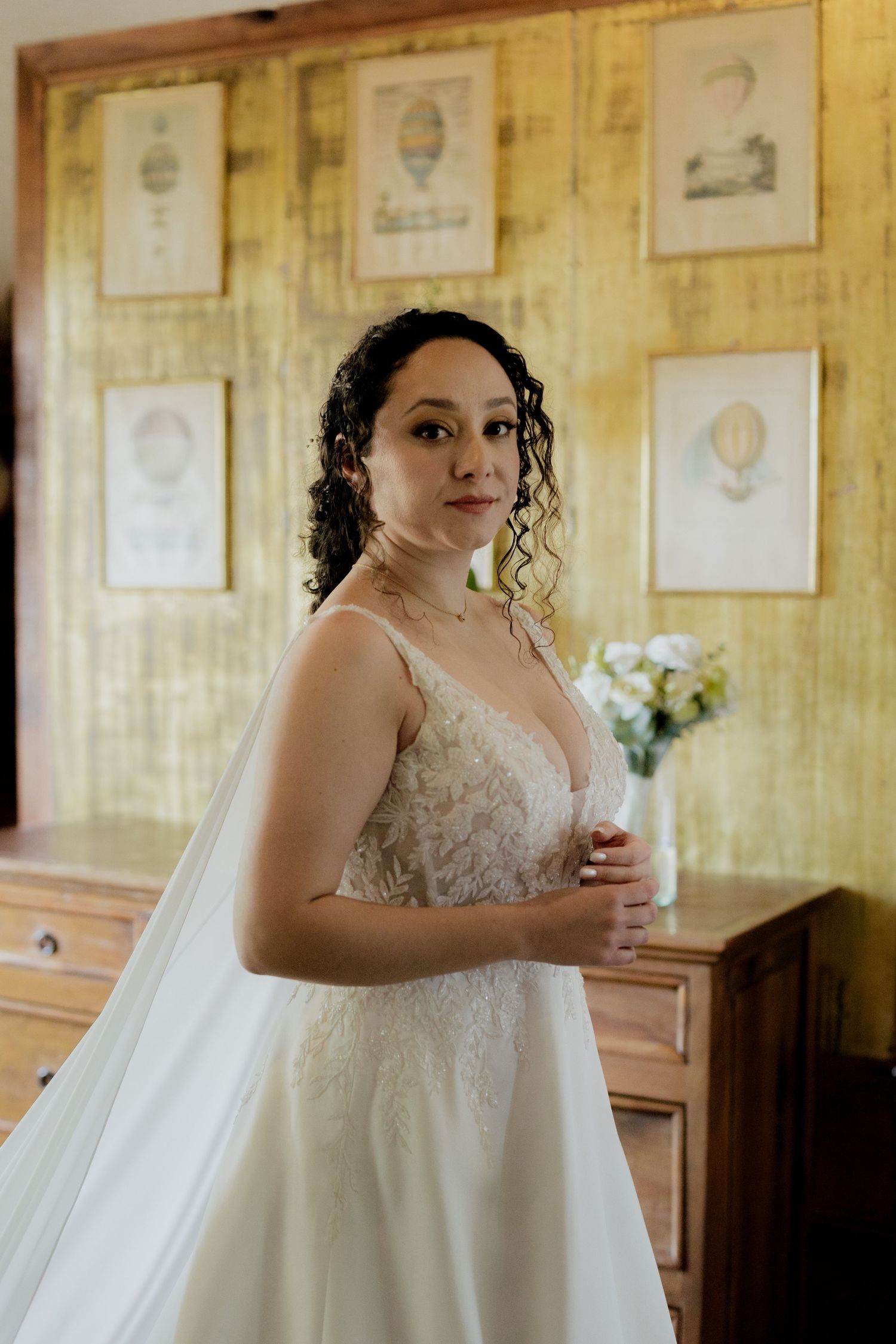 Bride in lace wedding gown poses in front of vintage wall with framed artwork.