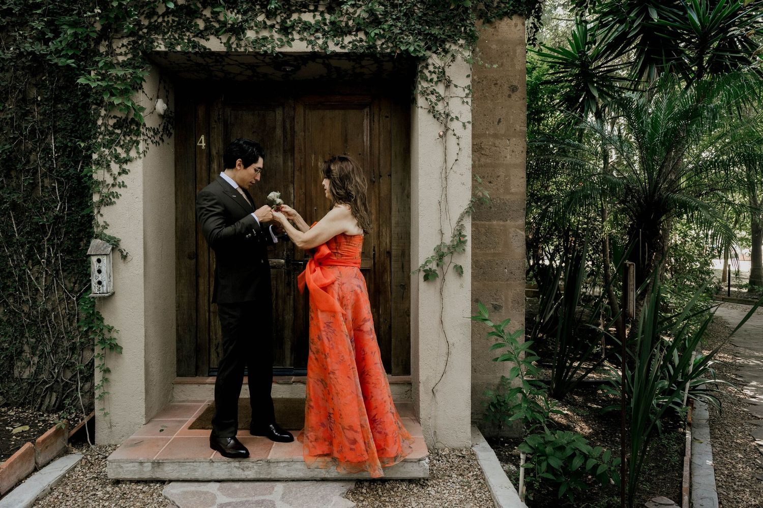 Garden pathway scene with figures in formal wear and orange dress against stone architecture.