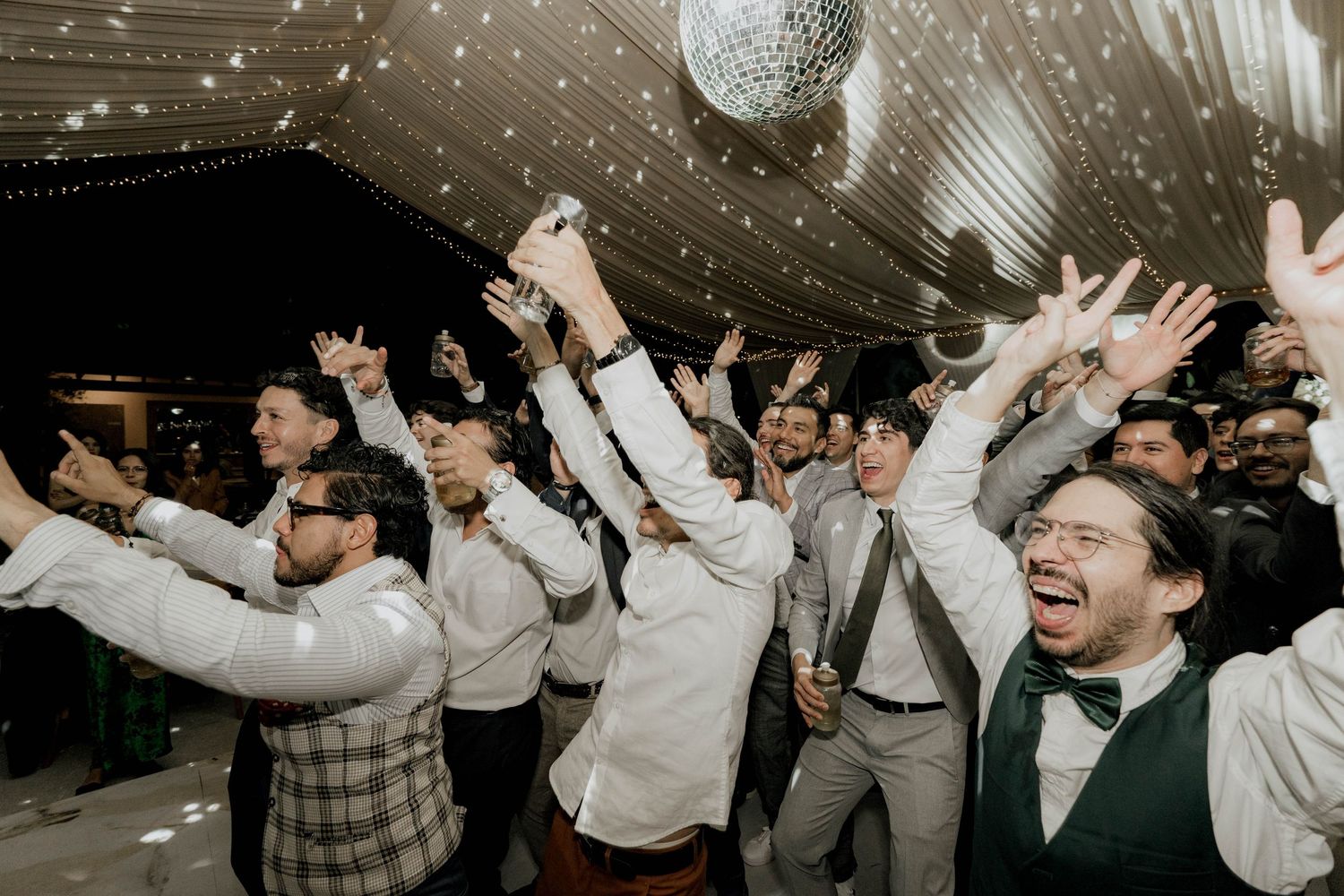 Wedding reception guests dancing and celebrating under disco balls and string lights in a tent venue.