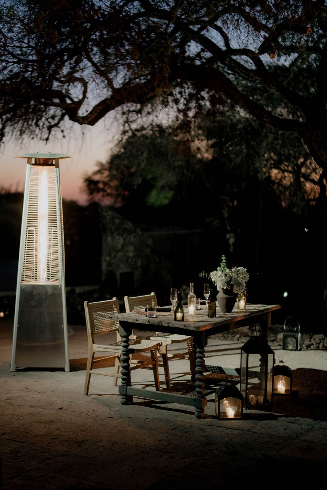 Outdoor dining setup with tall heater, lanterns and wooden table at dusk.