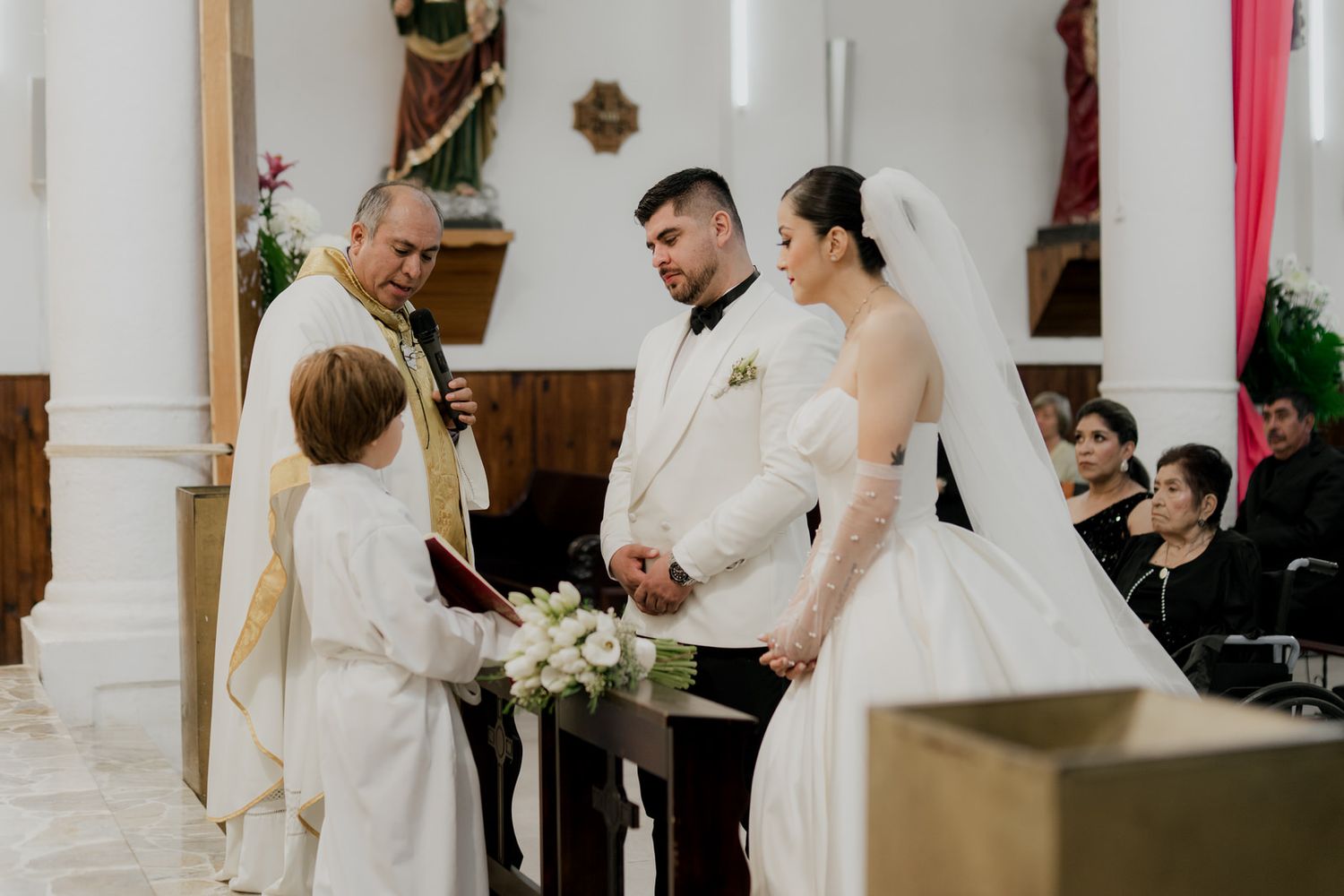 Wedding ceremony in a bright white church with traditional wooden pews and red accents.