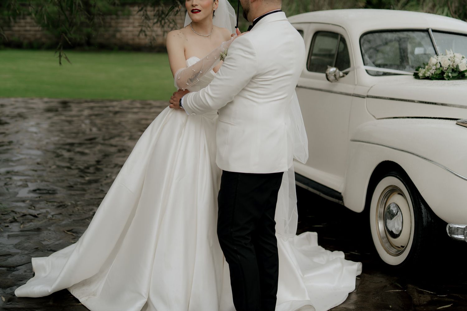 Couple in wedding attire poses next to a classic white vintage car outdoors.