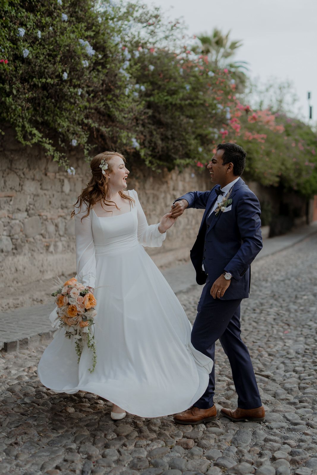 A joyful wedding couple dances on a cobblestone street surrounded by flowering trees.