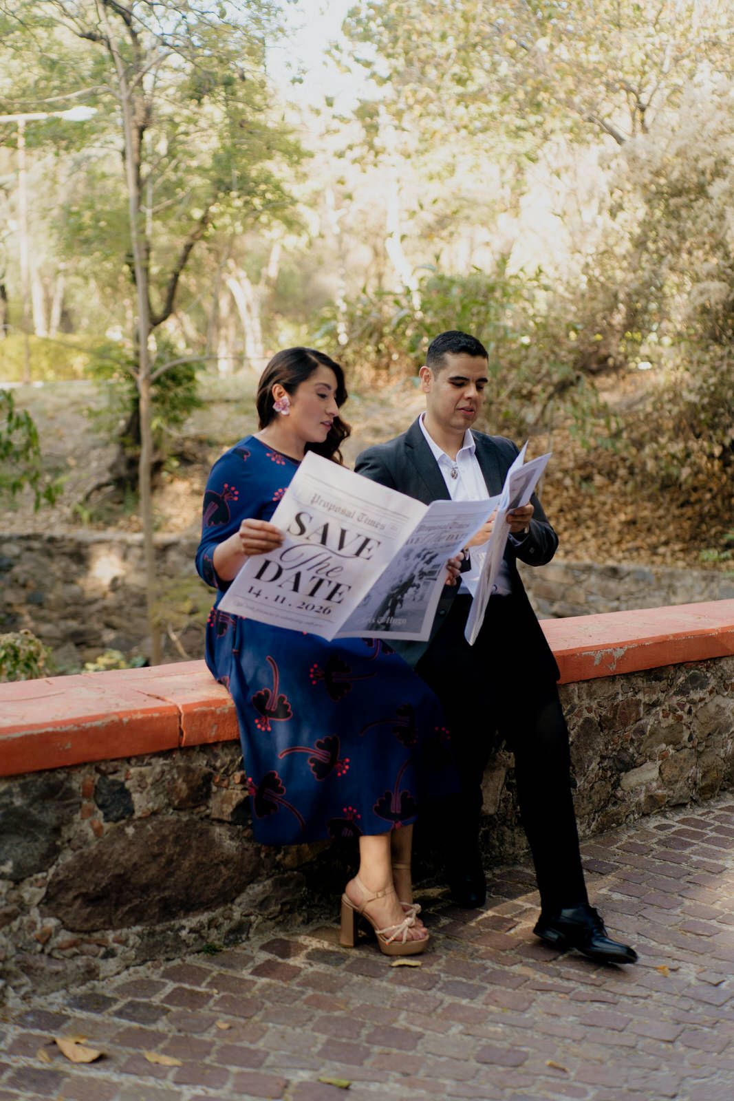 A couple sits on a brick wall reading newspapers during an engagement photoshoot.