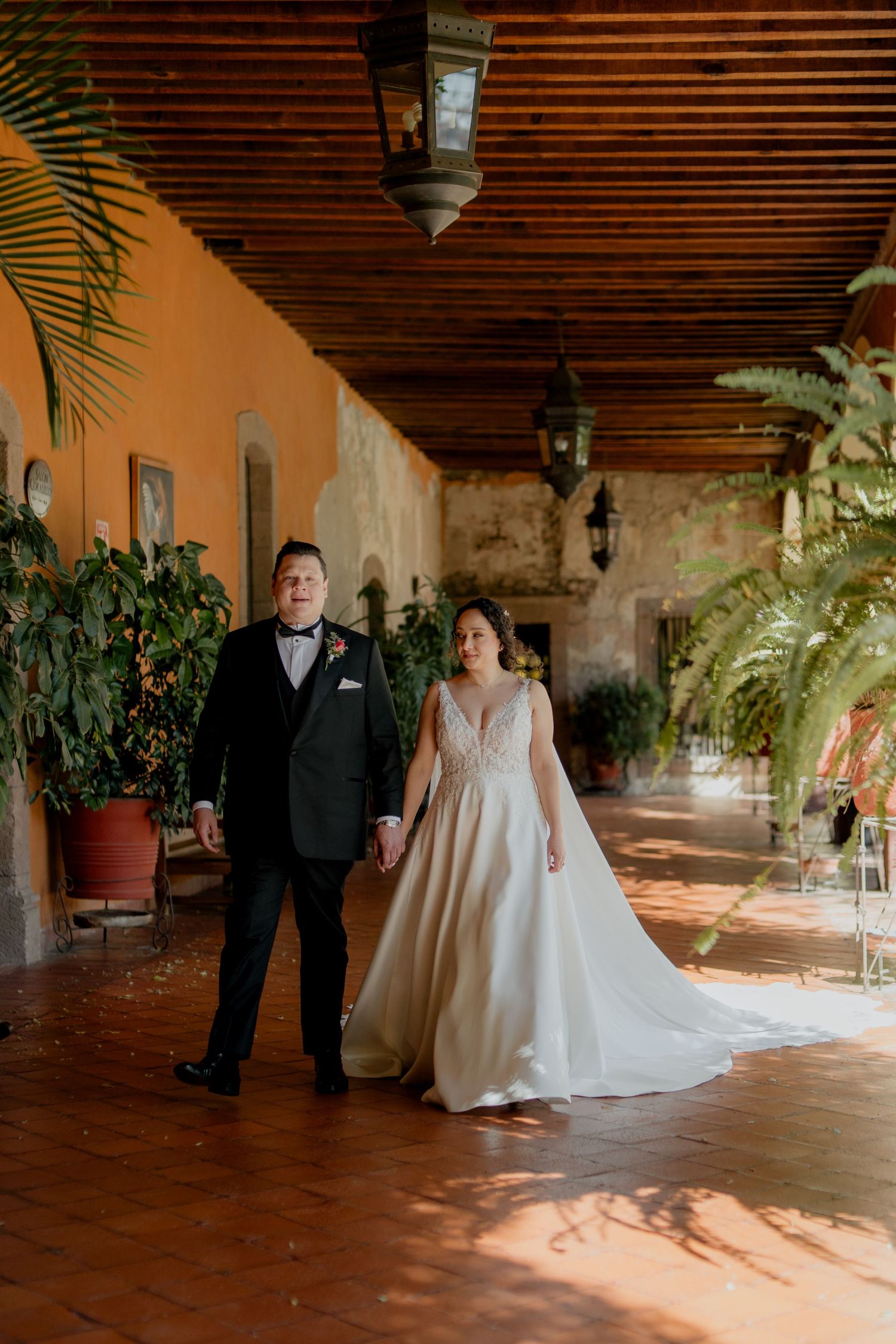 Wedding couple walks through a Spanish-style corridor with wooden beams and potted plants.