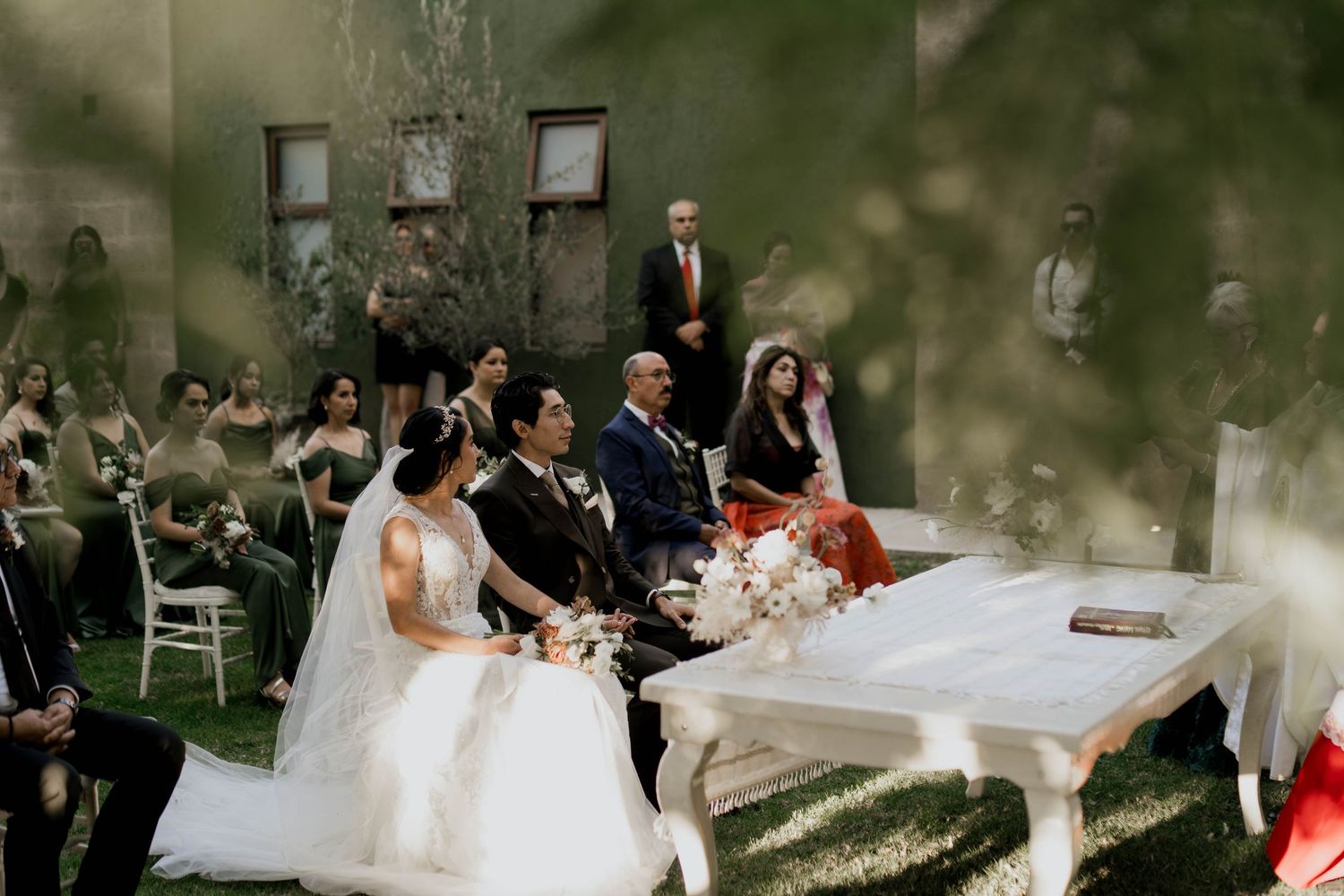 Wedding guests gather outdoors for a ceremony under trees with white chairs arranged in rows.