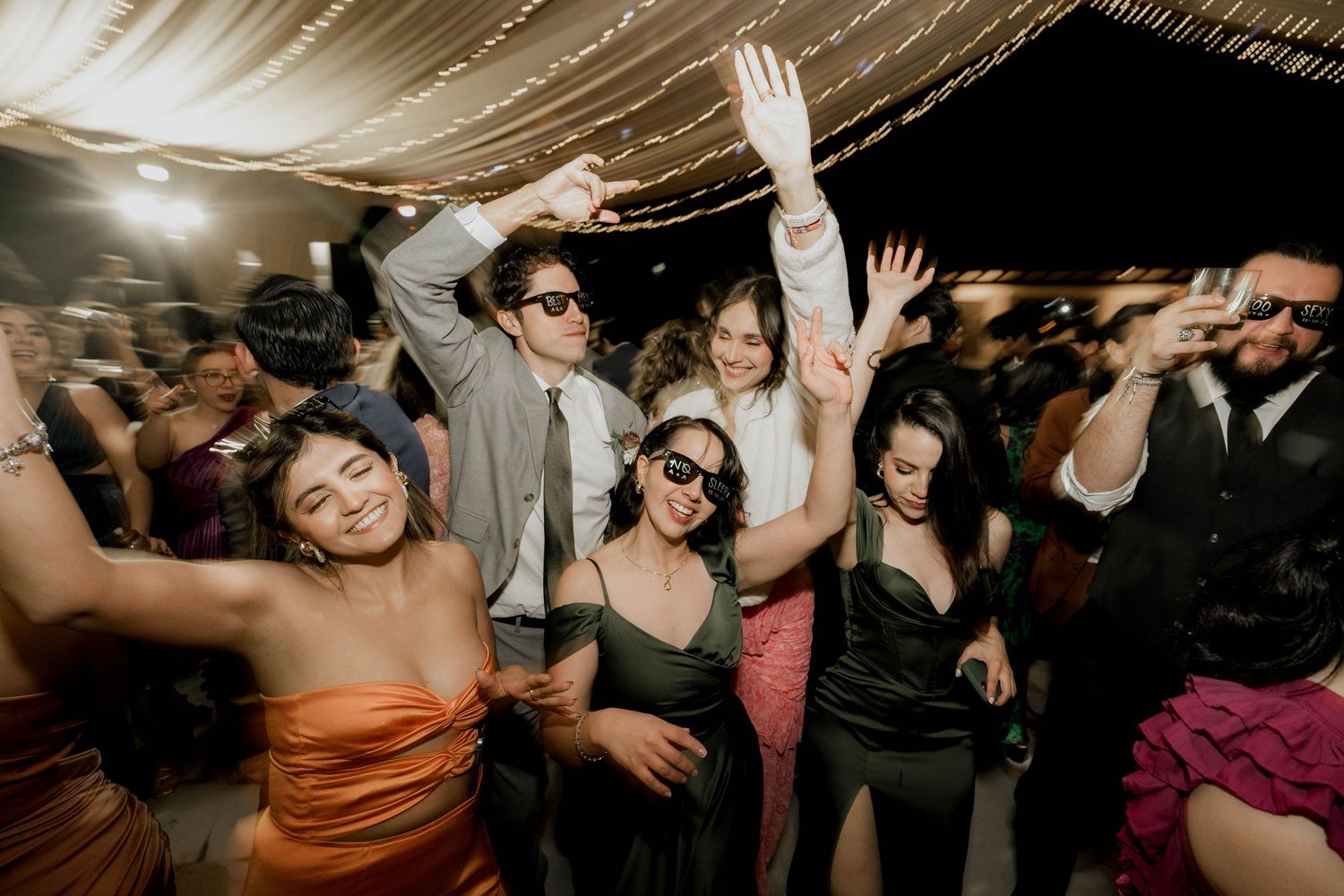 Energetic wedding guests dancing and raising hands on crowded dance floor under draped ceiling.
