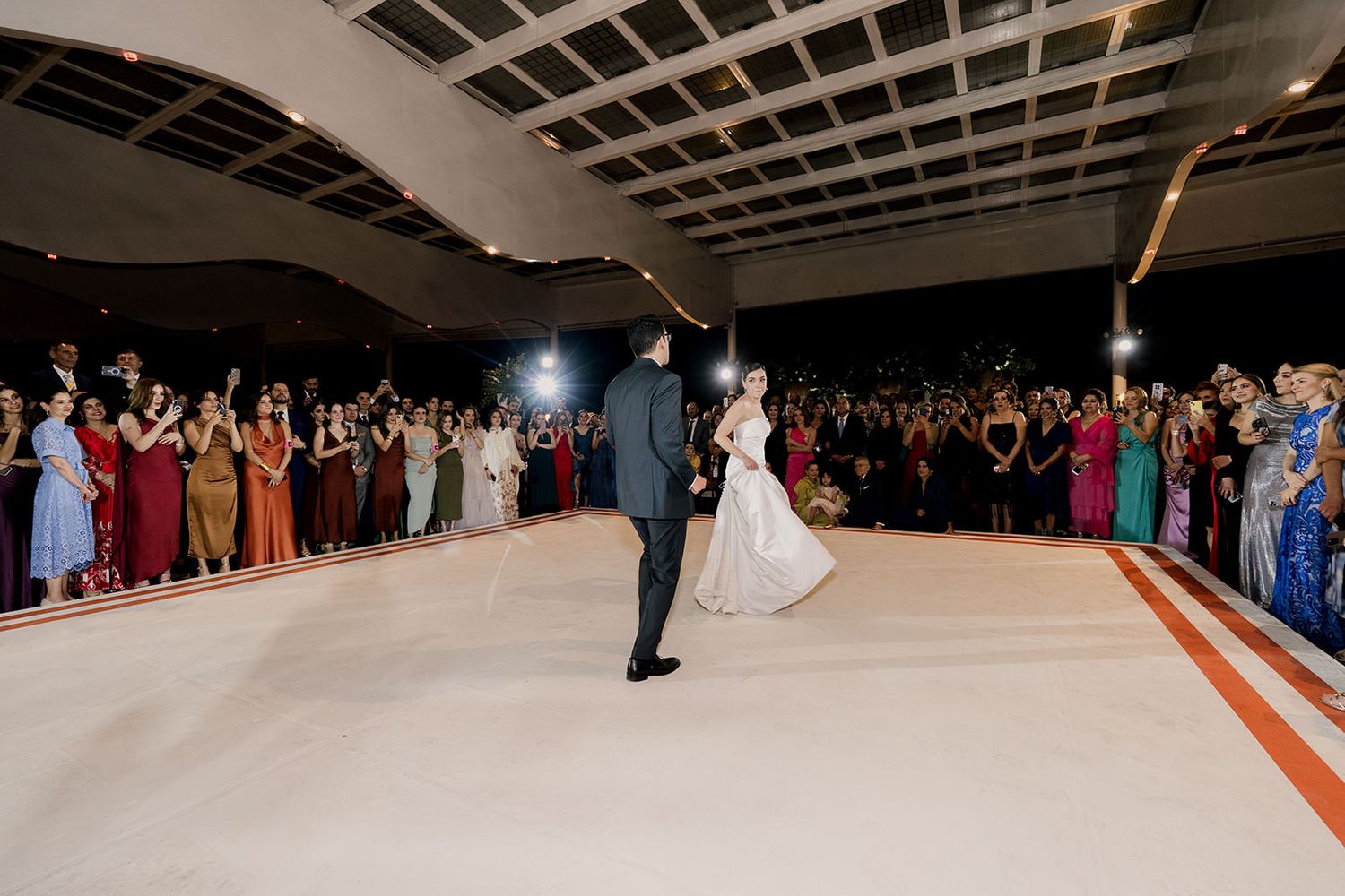 Bride and groom walk hand in hand on white dance floor surrounded by applauding wedding guests.