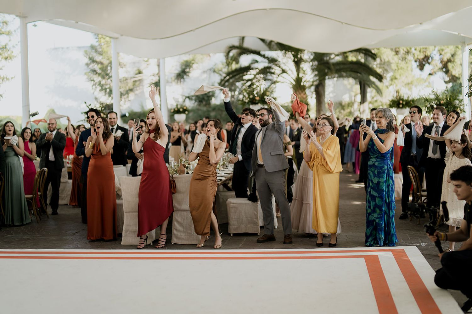 Wedding guests cheer and raise hands in joy during a lively outdoor reception under a white tent.