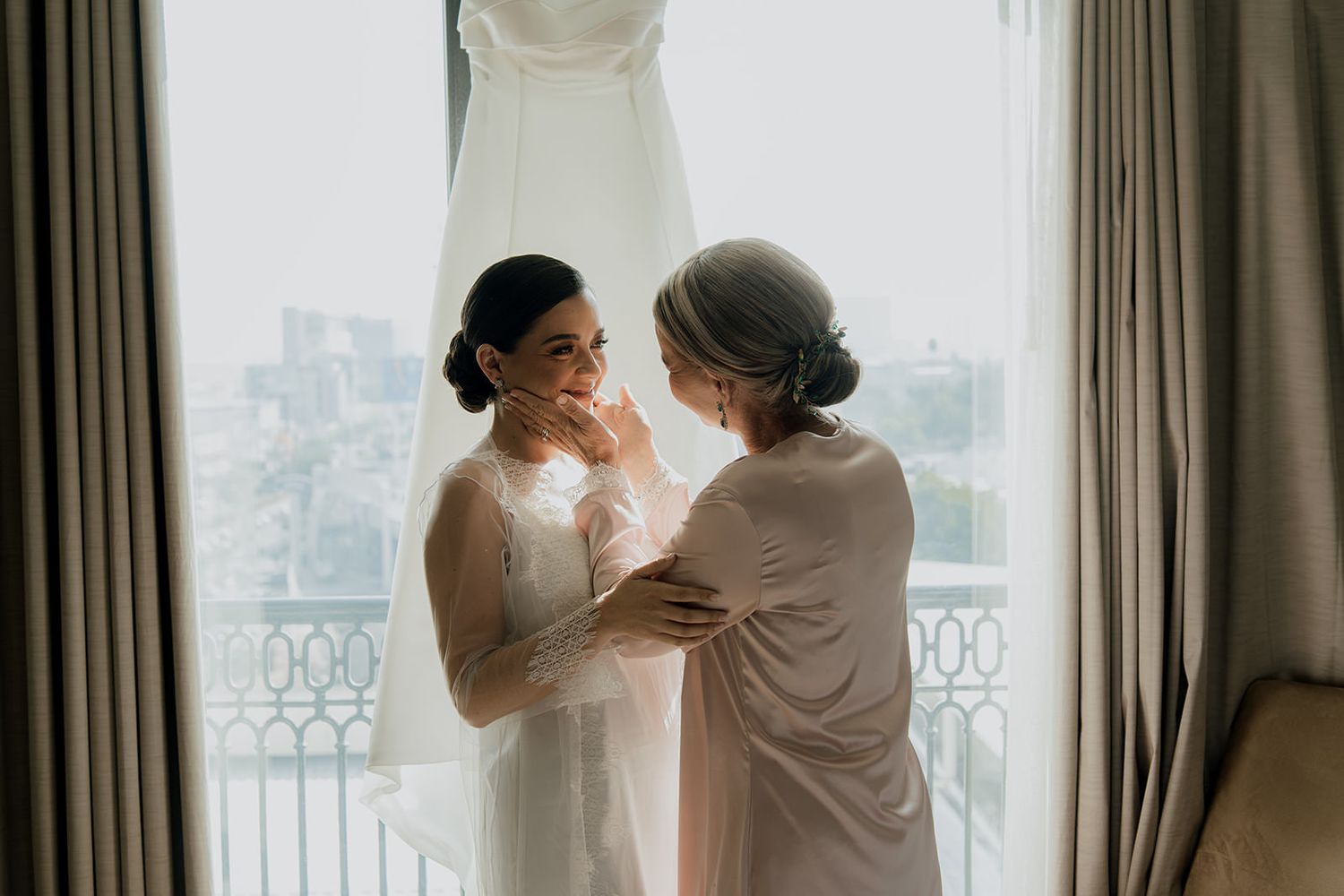 Emotional bride sharing tender moment with mother by window with wedding dress hanging behind.