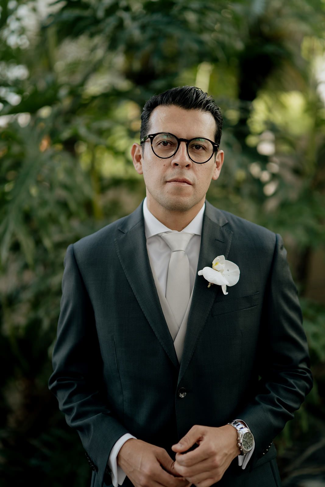 Dapper groom in dark suit with white boutonniere and glasses poses outdoors among lush green foliage.