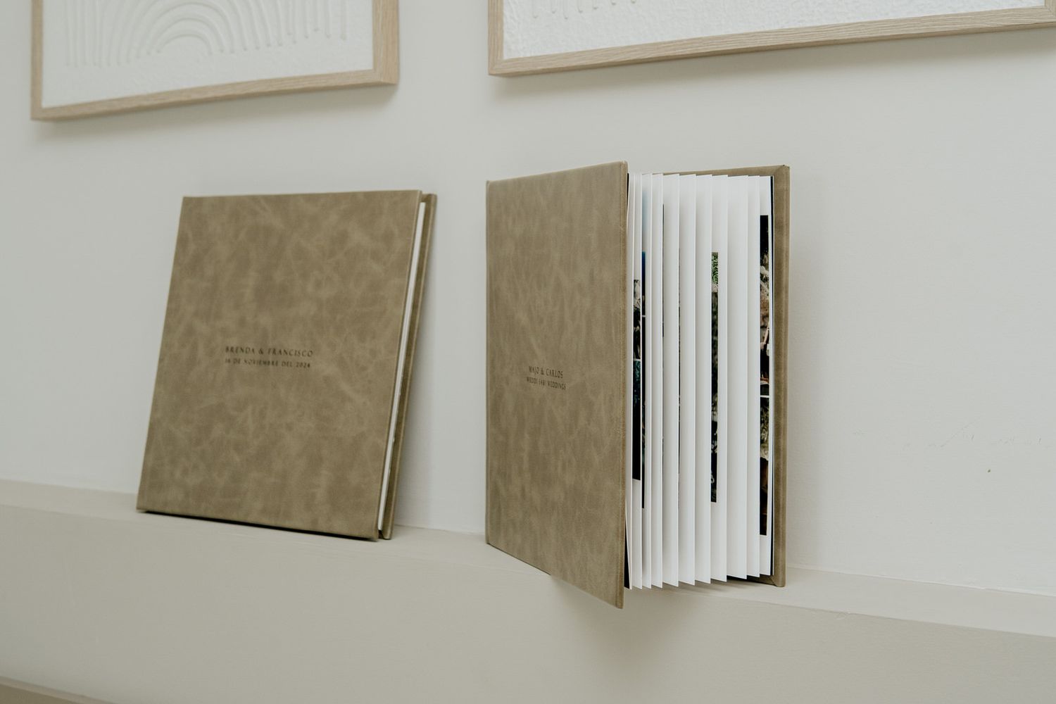 Two tan hardcover artist books displayed on a white wall, one open showing layered pages.