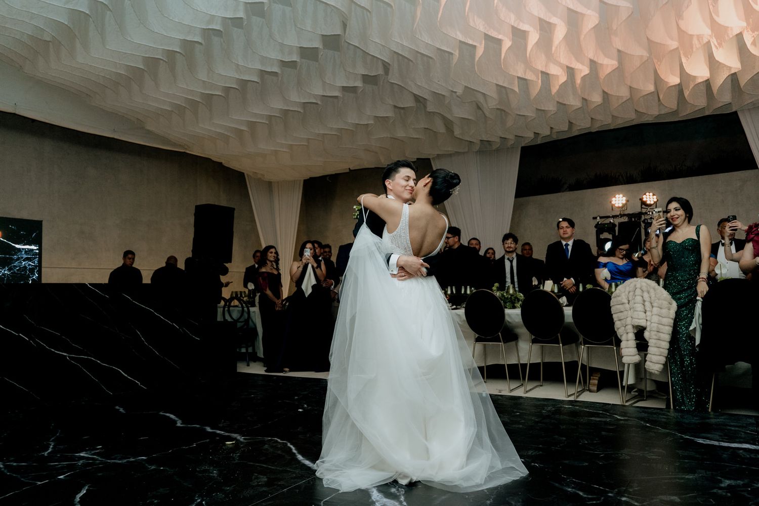 Bride and groom share romantic first dance kiss in elegant venue with geometric ceiling and wedding guests seated nearby.