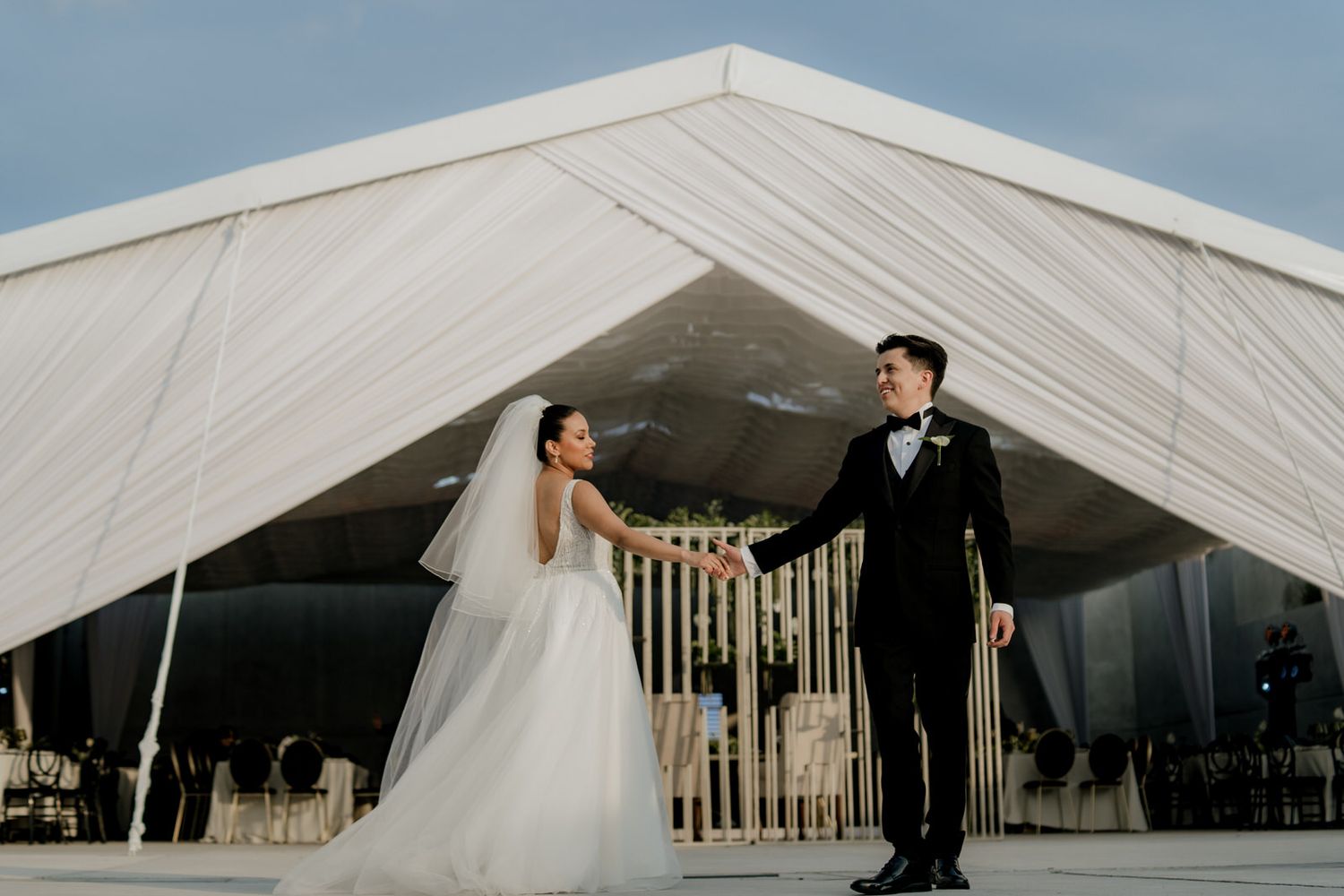 Bride and groom hold hands outside elegant white draped wedding tent, bride in tulle gown, groom in black tuxedo.