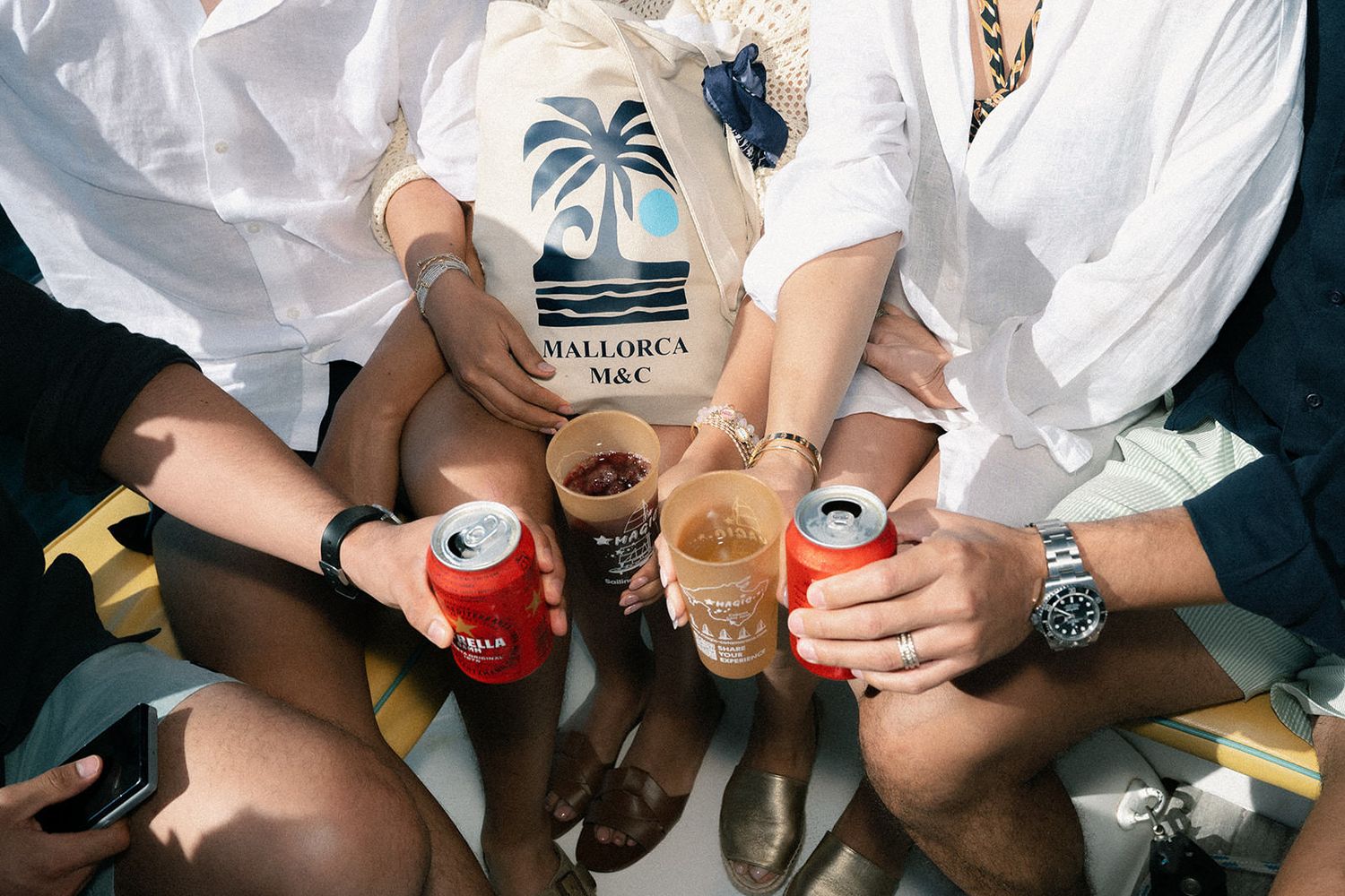 Friends holding beverages with Mallorca Bay branded tote bag visible in casual summer gathering.