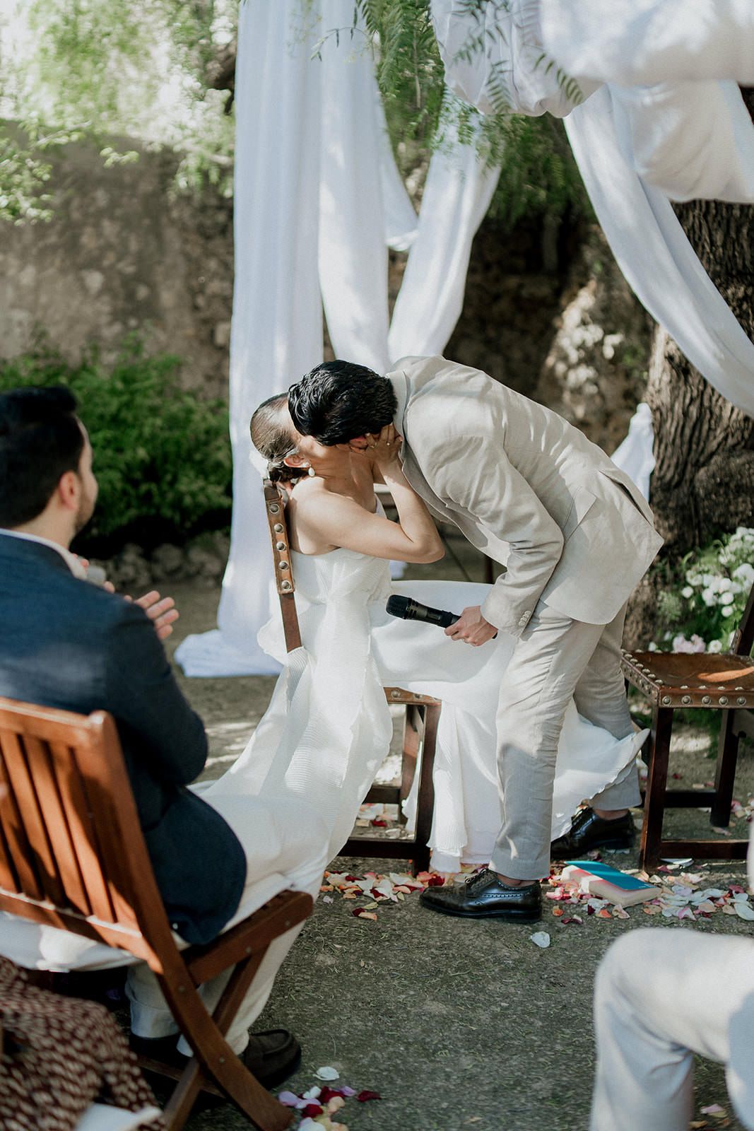A couple shares a tender moment during an outdoor wedding ceremony under flowing white fabric and natural greenery.