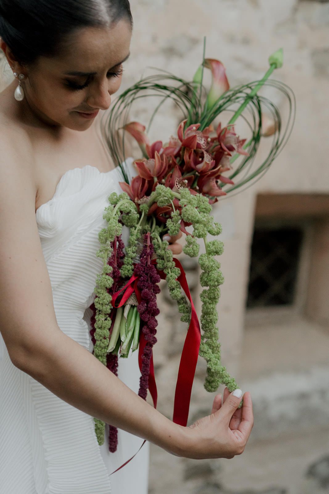 A wedding bouquet featuring burgundy flowers and green bells tied with red ribbon is held against a white dress.