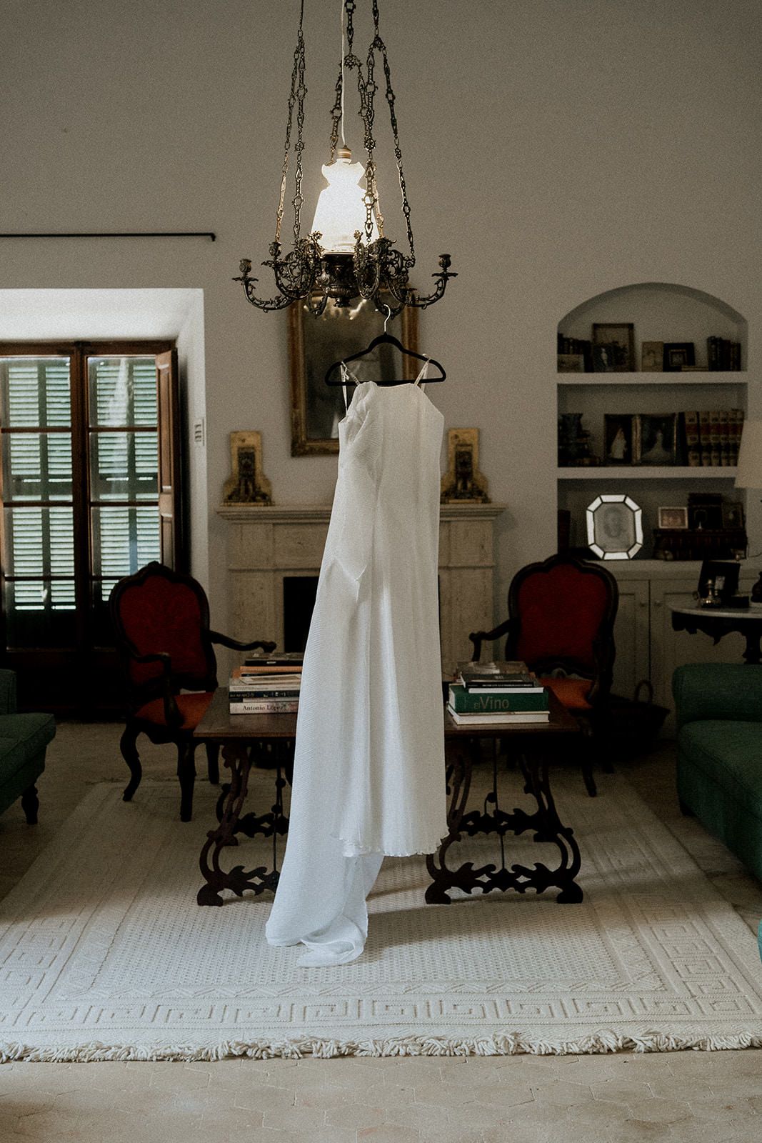 Elegant white wedding dress hangs from an ornate chandelier in a vintage-style room with wooden floors.