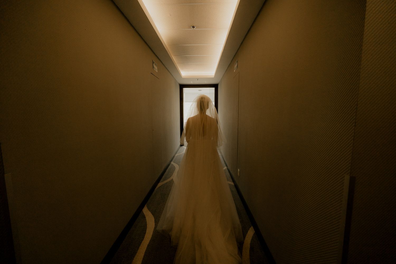 Bride in a white wedding gown standing at the end of a dimly lit hallway, captured from behind.