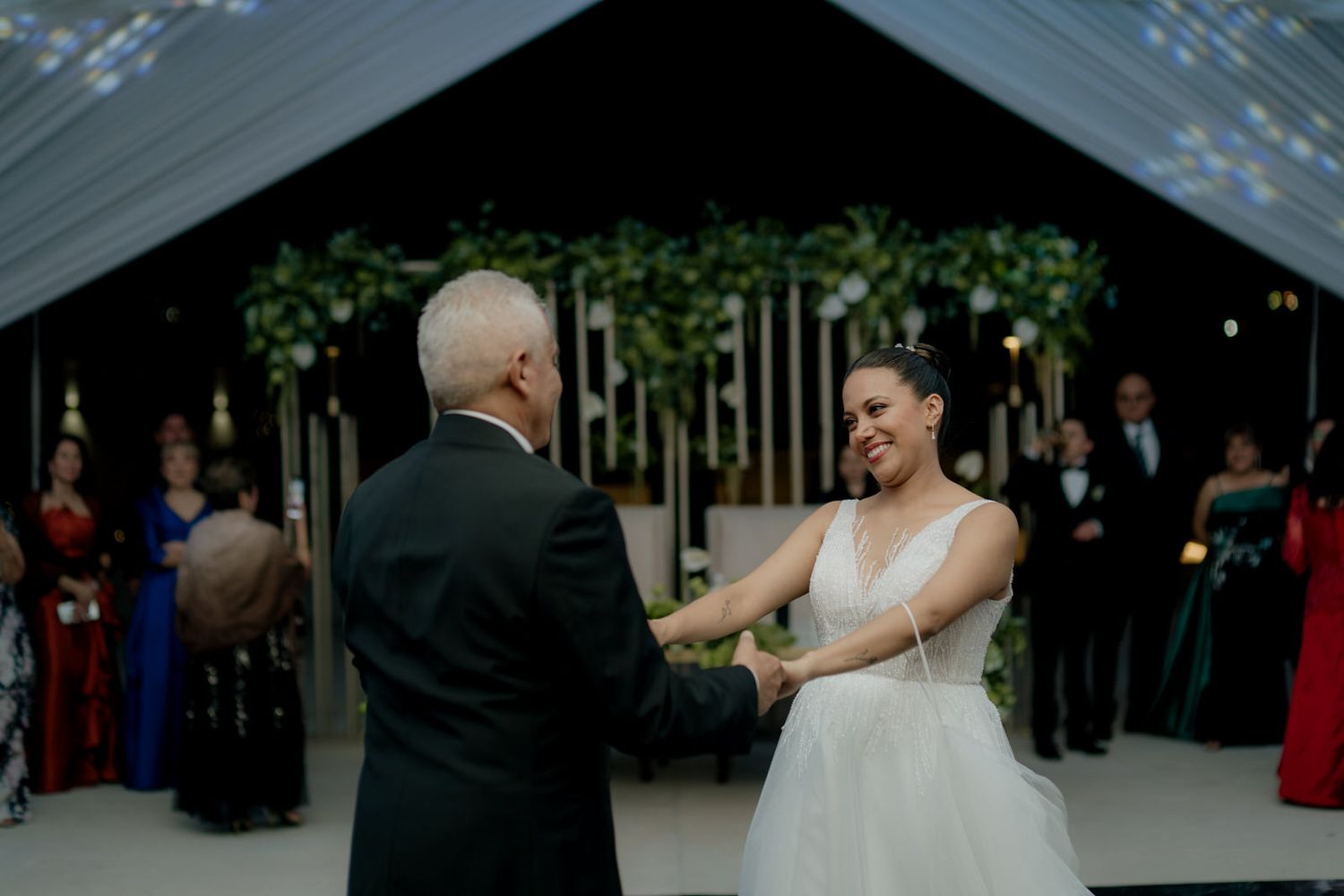 Bride in white gown smiles while sharing a dance with older gentleman at elegant wedding reception under draped tent.