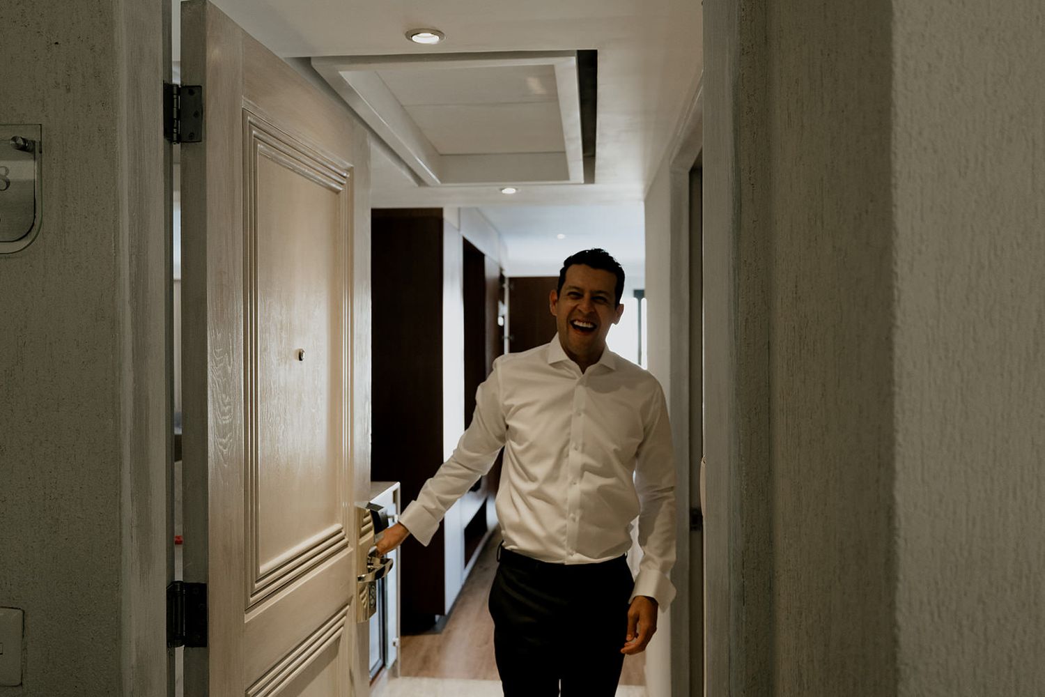 Smiling groom in a white dress shirt walking through a hotel corridor on his wedding day.