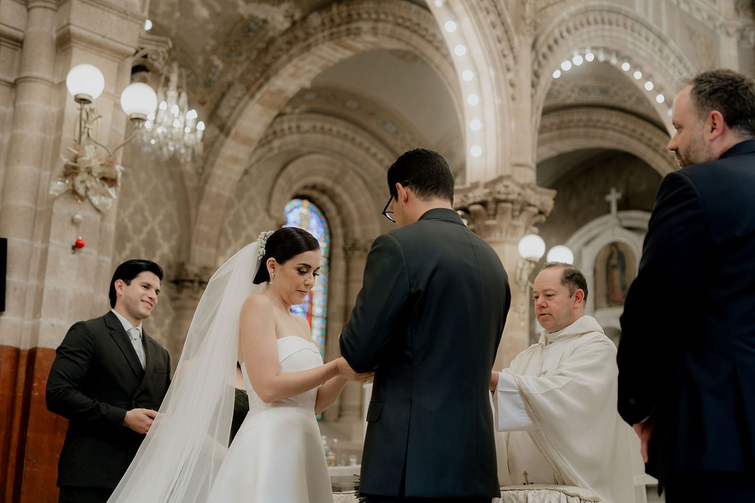 Bride and groom exchange vows at elegant church wedding ceremony with priest and witness present.