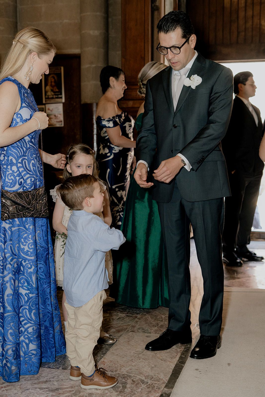 Dapper groom in dark suit greets young children with guests in formal attire at wedding venue.
