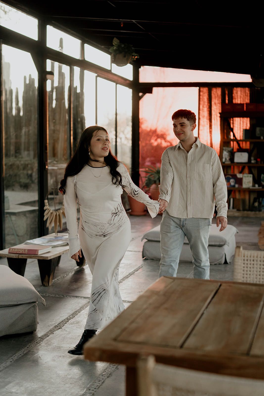 Two people in white and light colored clothing standing near wooden tables indoors.