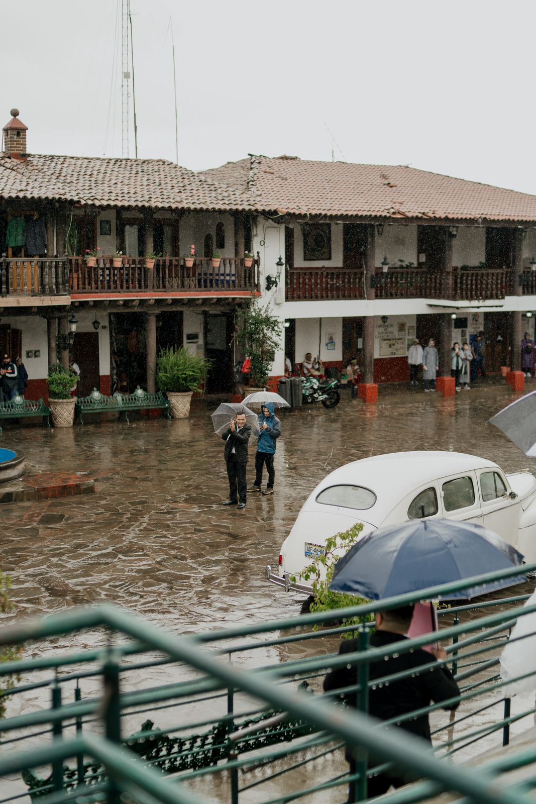 Vintage car parked in colonial courtyard during rainfall with people under umbrellas.