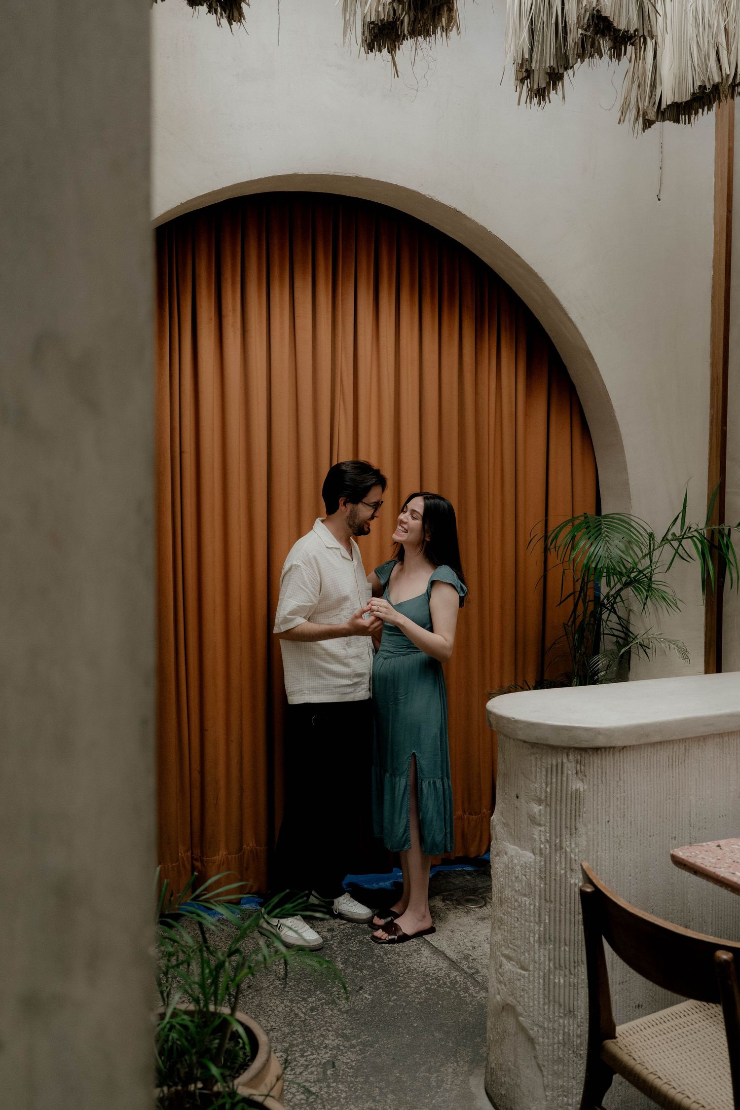 A couple shares a tender moment in front of an arched doorway with orange curtains.