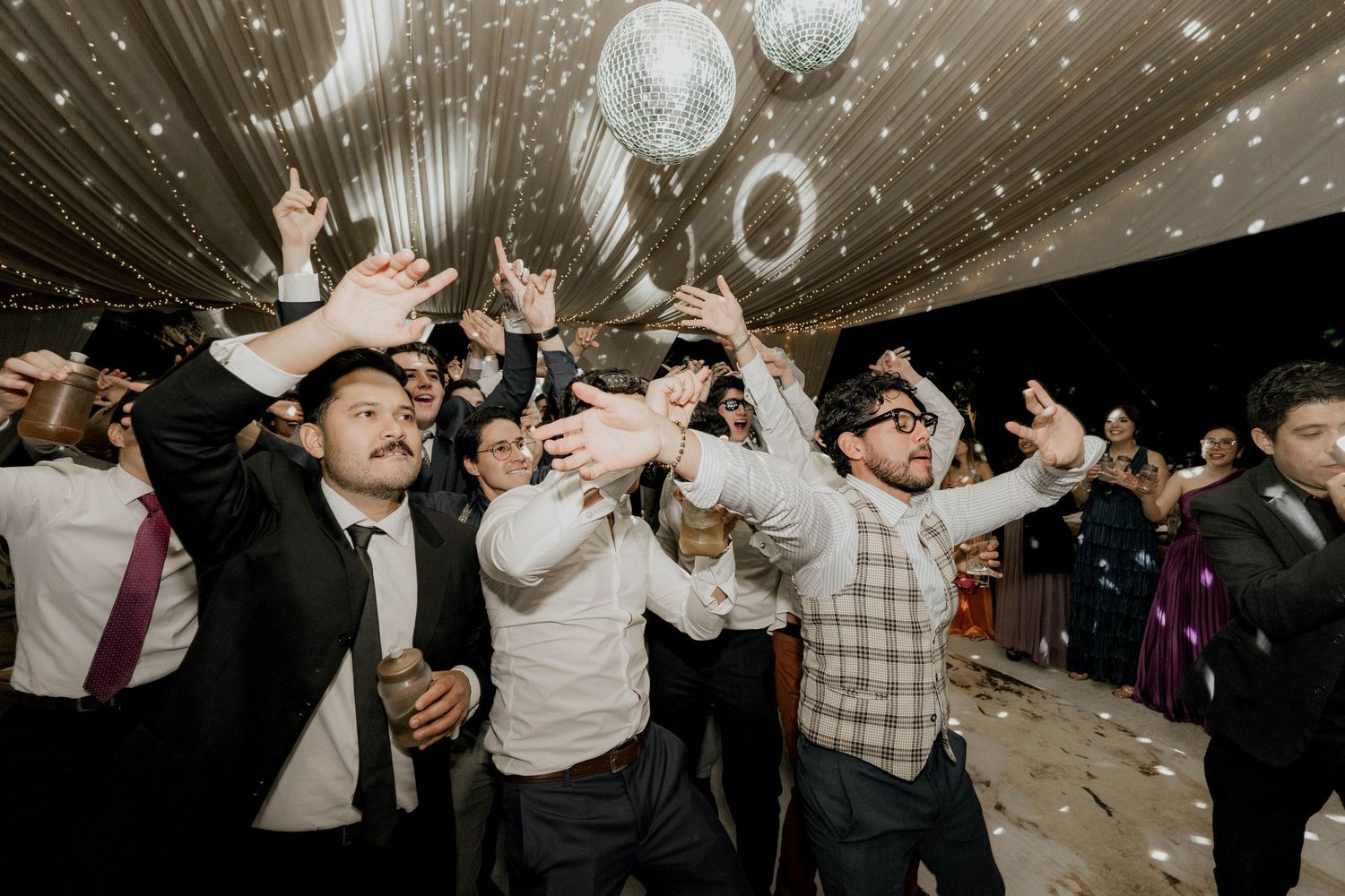 Party guests celebrating with hands in the air beneath sparkling disco balls and string lighting.