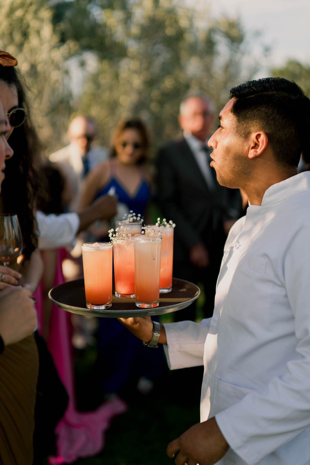 Servers carrying trays of pink cocktails at an outdoor wedding reception.