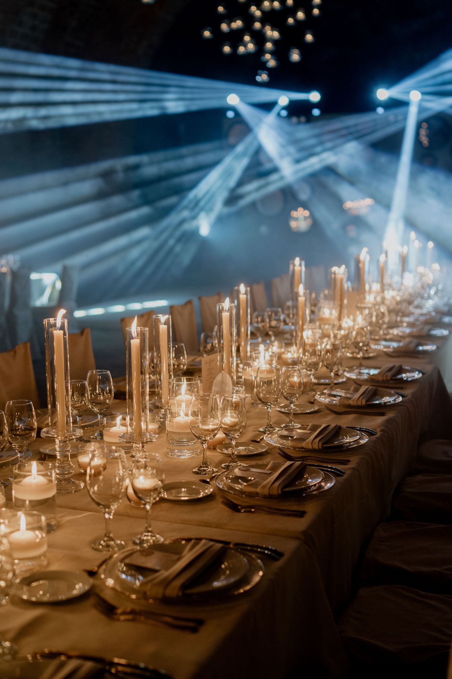Elegant table settings with glowing candles and crystal glassware under dramatic blue lighting.