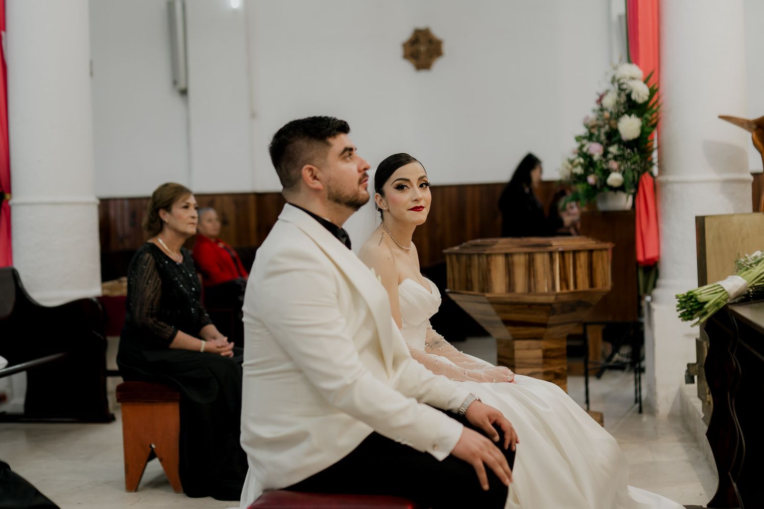 A couple in white formal attire sits attentively during their church wedding ceremony.