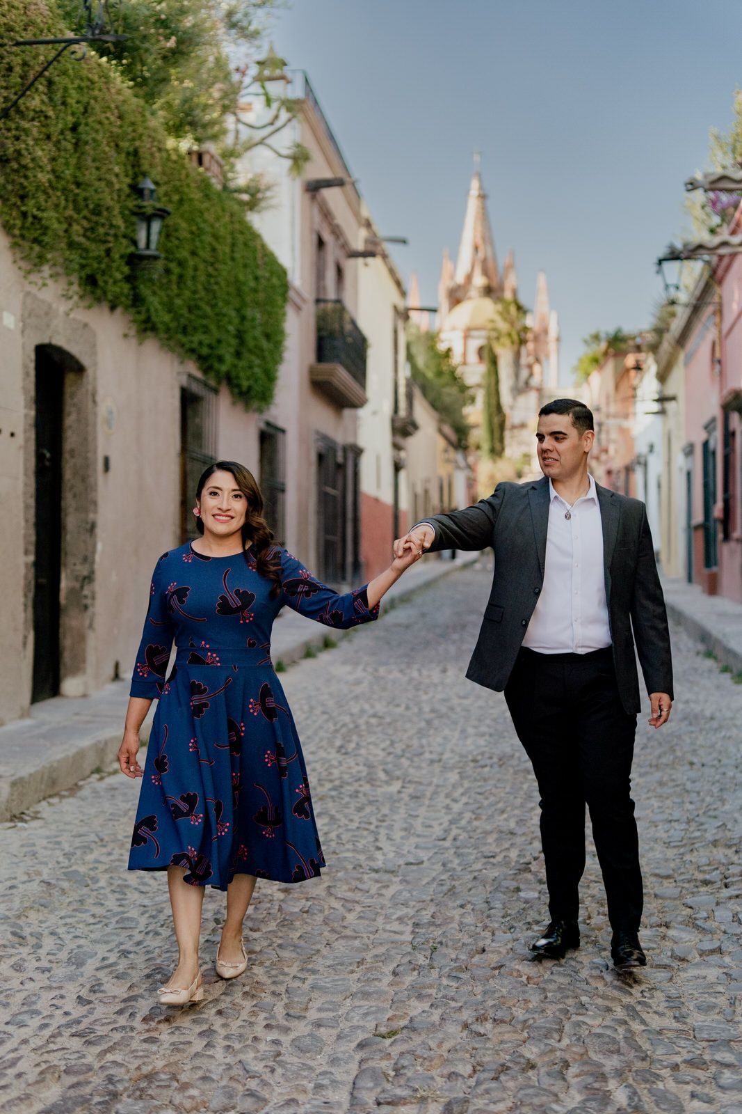 A couple walks hand in hand down a cobblestone street with a church spire visible in the background.