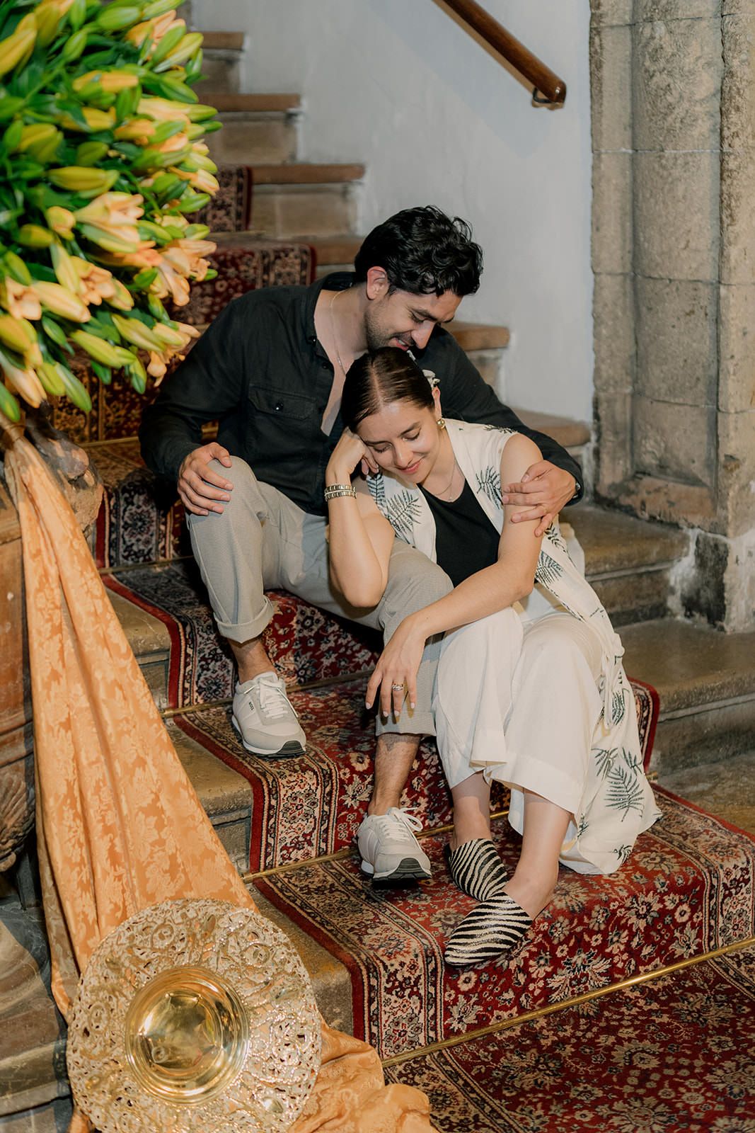 Two people share an intimate moment sitting on ornate carpeted stairs beside yellow lilies and decorative elements.