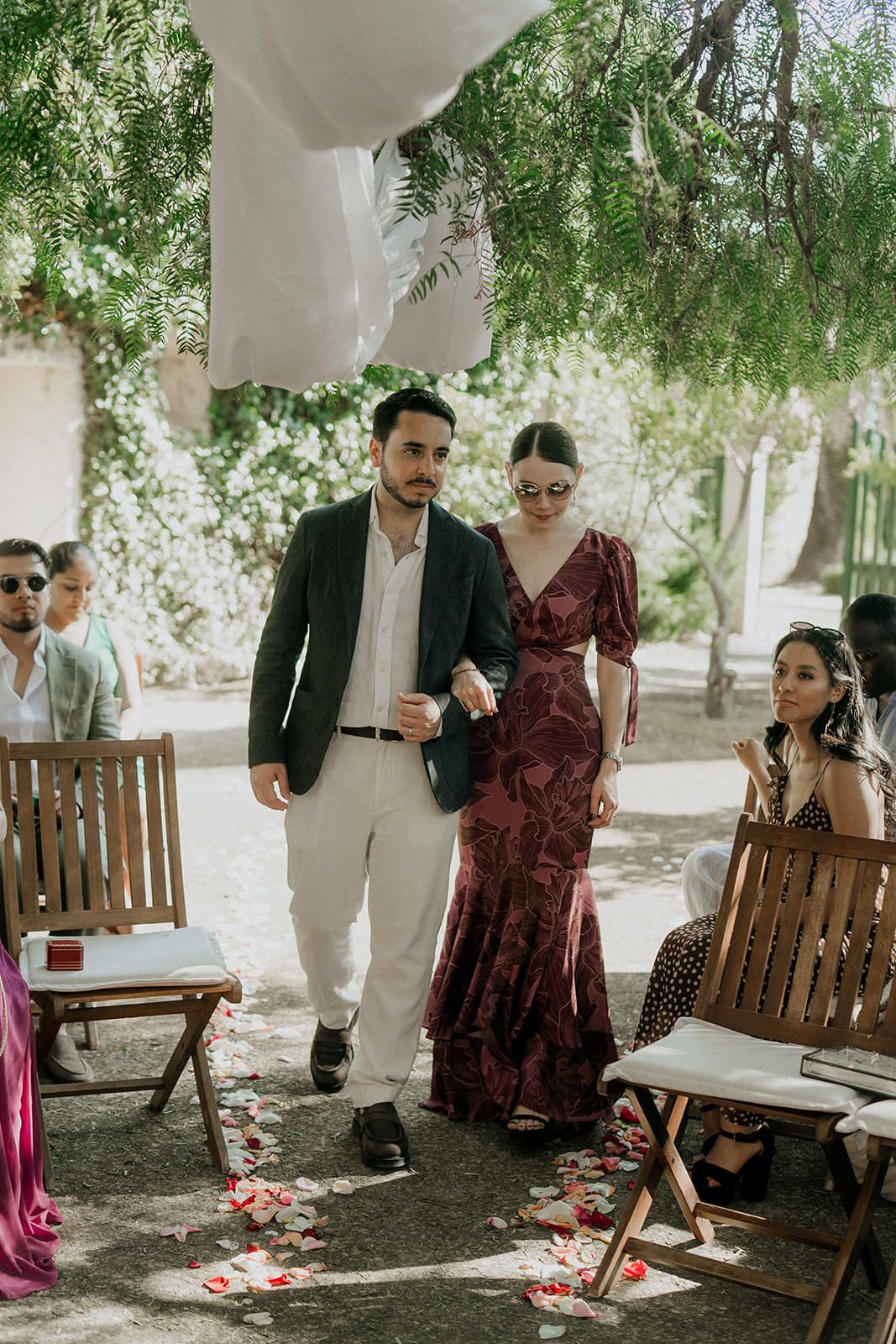 A romantic outdoor gathering shows couples seated at wooden benches under shady trees during a casual celebration.