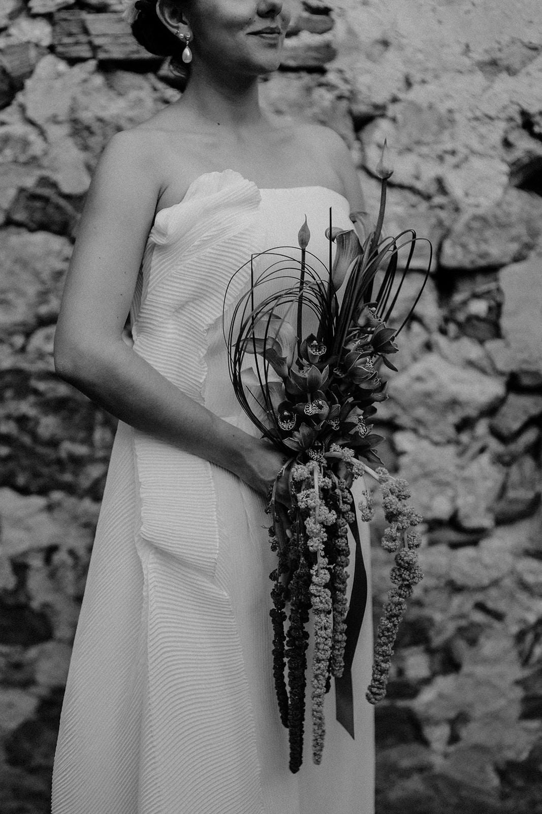 Black and white close-up of a bridal bouquet with cascading flowers against a stone wall.