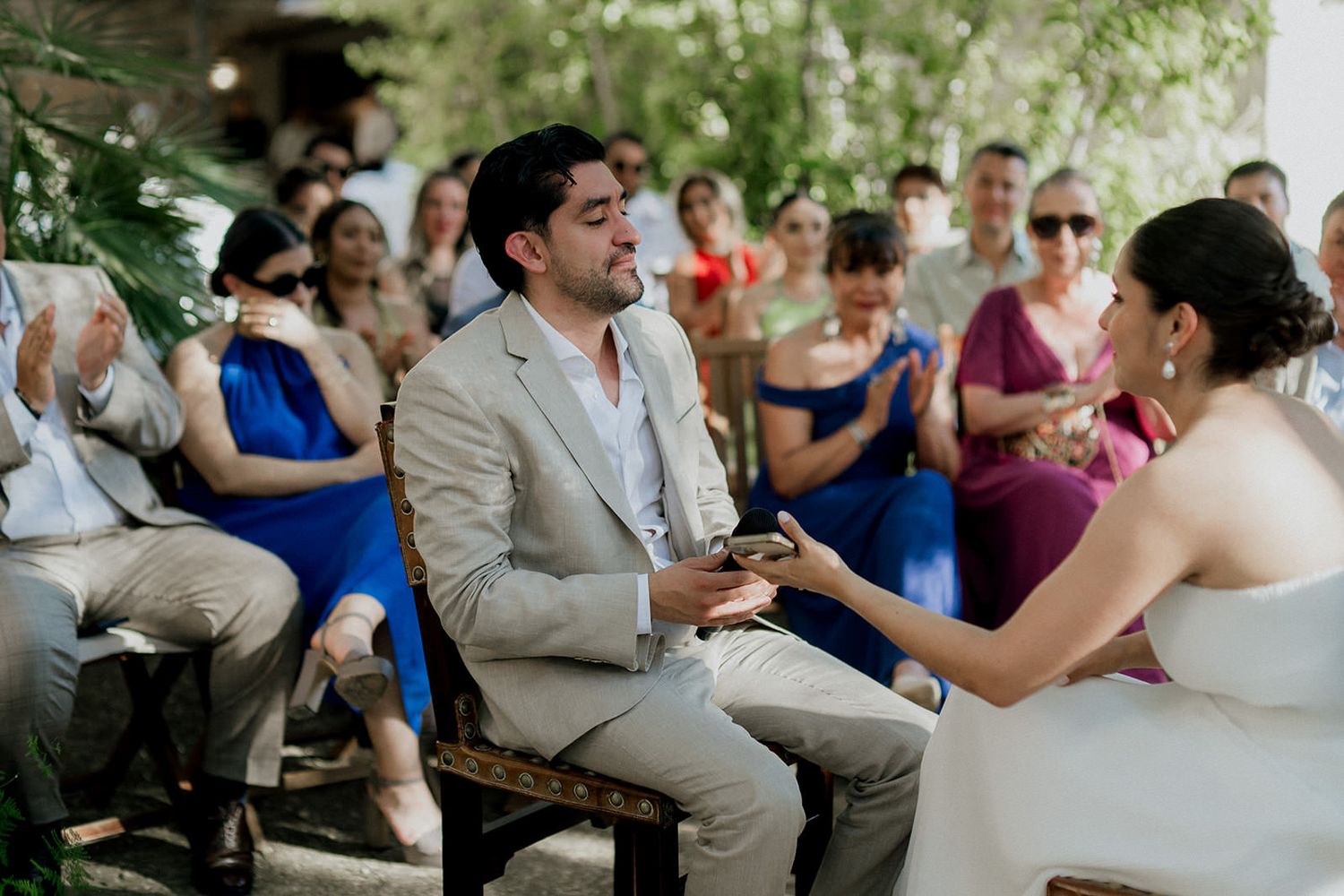 Wedding guests gather outdoors under trees for a ceremony with people in blue and beige attire.